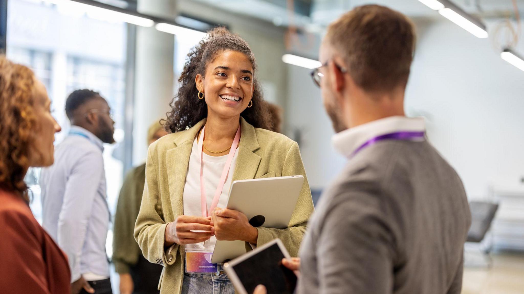 A woman with brown hair wearing a green blazer chats to a grey-haired man with grey glasses. A woman with ginger hair and a red blazer stands beside them