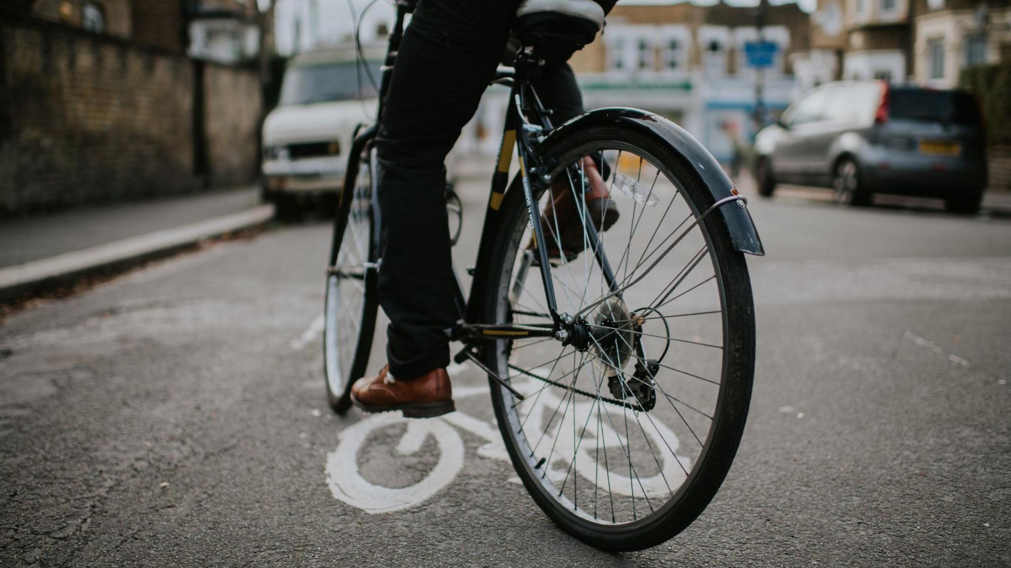 A person riding a bicycle, only their legs are visible in the shot. They are on a street with some cars visible