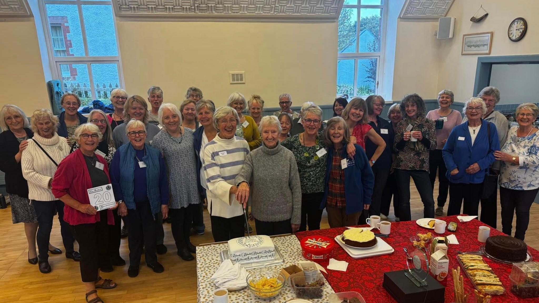 A group of around 30 women standing behind a table with cakes inside a village hall