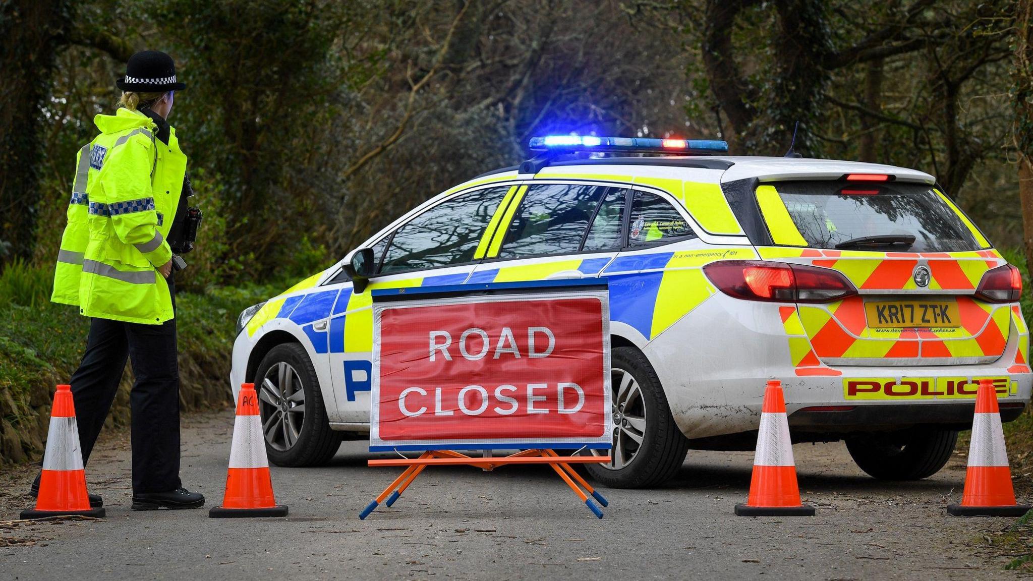 A stationary police car parked on a road has its blue lights flashing. There is a row of cones in front of it and a sign reading "ROAD CLOSED". A female police officer wearing uniform and a fluorescent jacket has her back to the camera. There are trees behind the car.