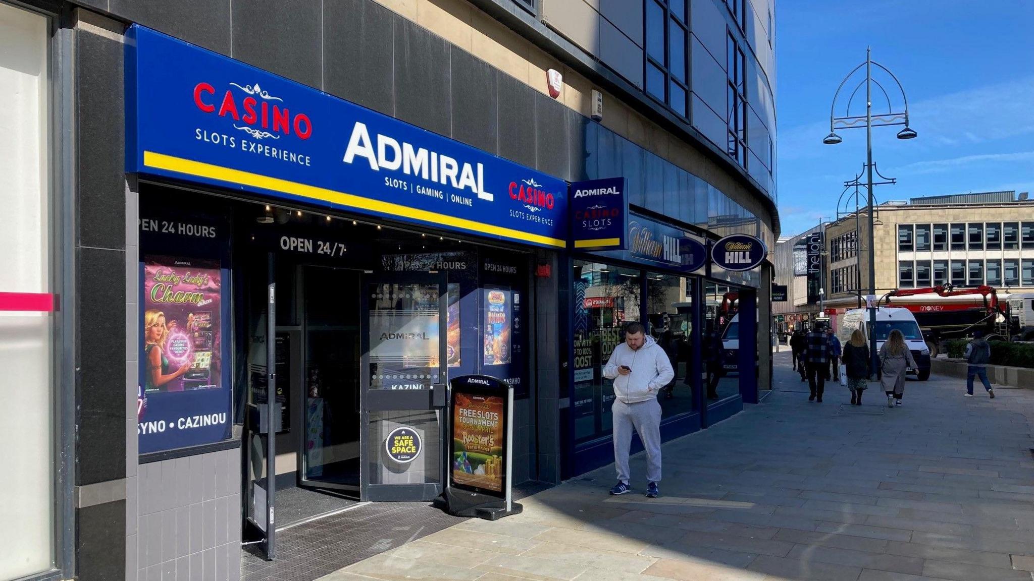 The shop front of a slot machine arcade with a man wearing a grey tracksuit looking at his mobile phone and walking past the entrance.