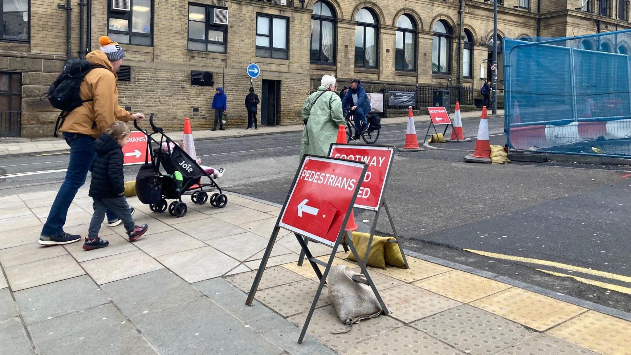 Red and white temporary signs directing pedestrians across a road junction with a fenced-off area to the right and traffic cones on the pavement.