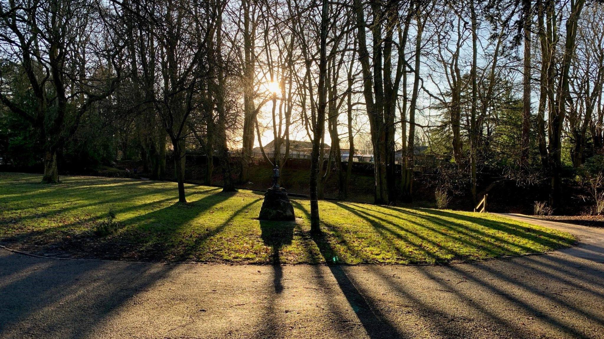 Light pours through the trees as the sun sets in a park: lines are drawn by shade that cross the grass verge and come towards the viewer. Statue almost entirely obscured by darkness in mid-ground