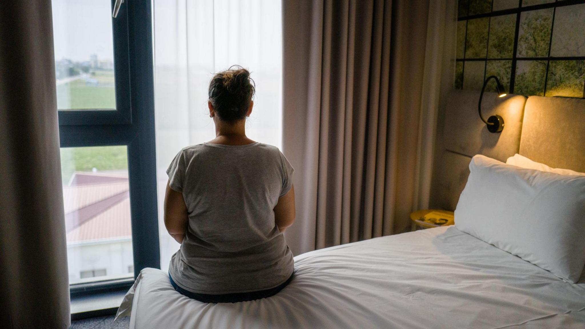 A woman sitting alone on a bed in a hotel room, looking out of the window.