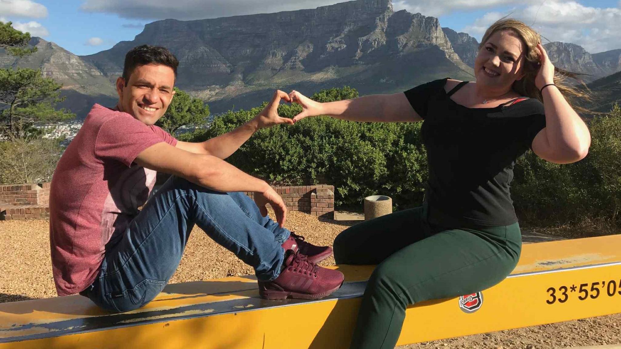 A man wearing a red t-shirt, jeans and red t-shirt, sits on a yellow bar in the air, holding his hand in a love heart shape. Next to him a woman with long flowing hair, holds her hair back and also puts her hand into a heart shape next to his, sitting next to him on a bar. There are mountains behind them on a sunny day.