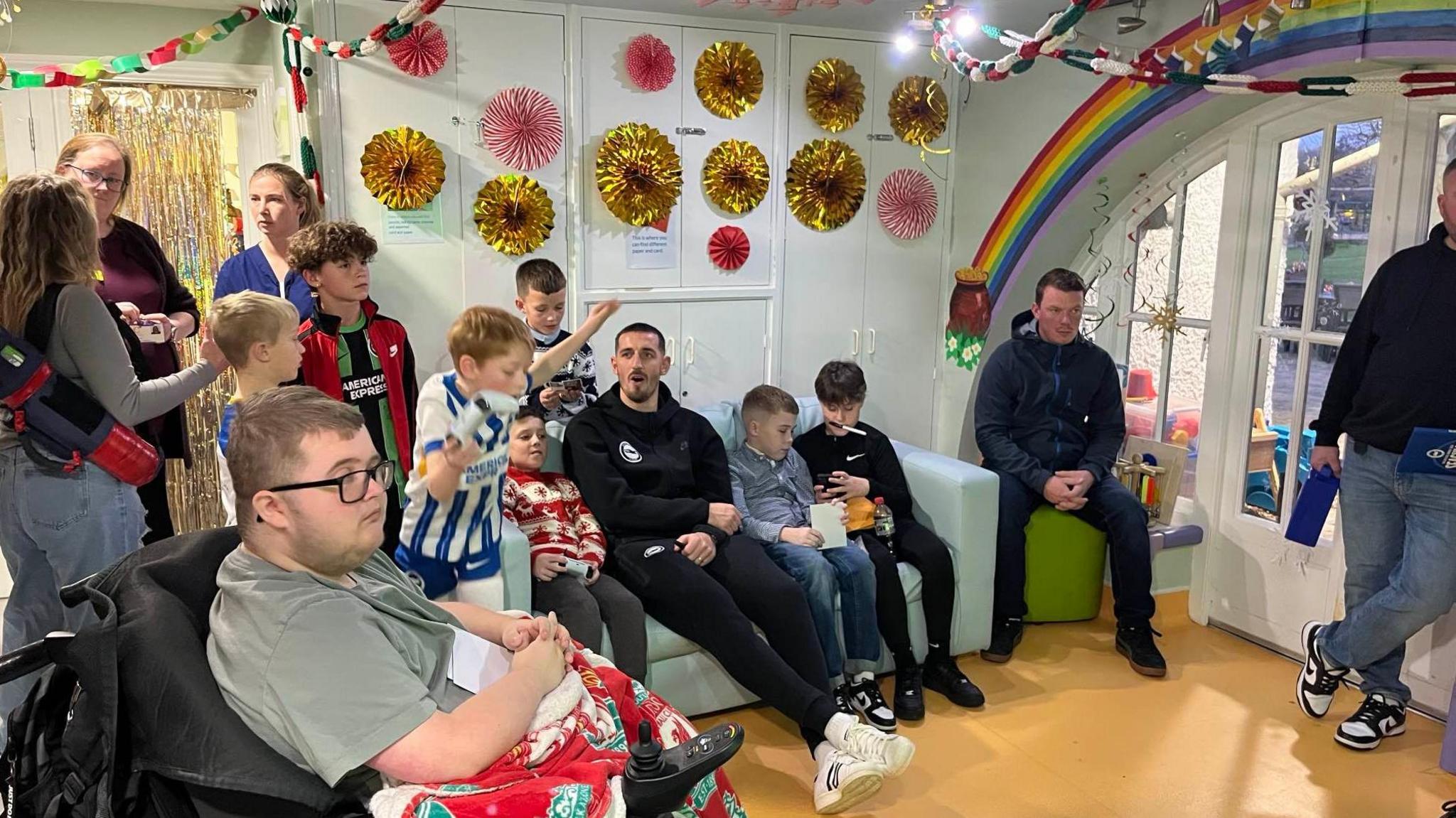 A group of adults and children, some sitting, some in wheelchairs, some standing, gather around a games console and TV. One child wearing a football kit is waving his arms in celebration. Christmas decorations can be seen on the ceiling and in the background.