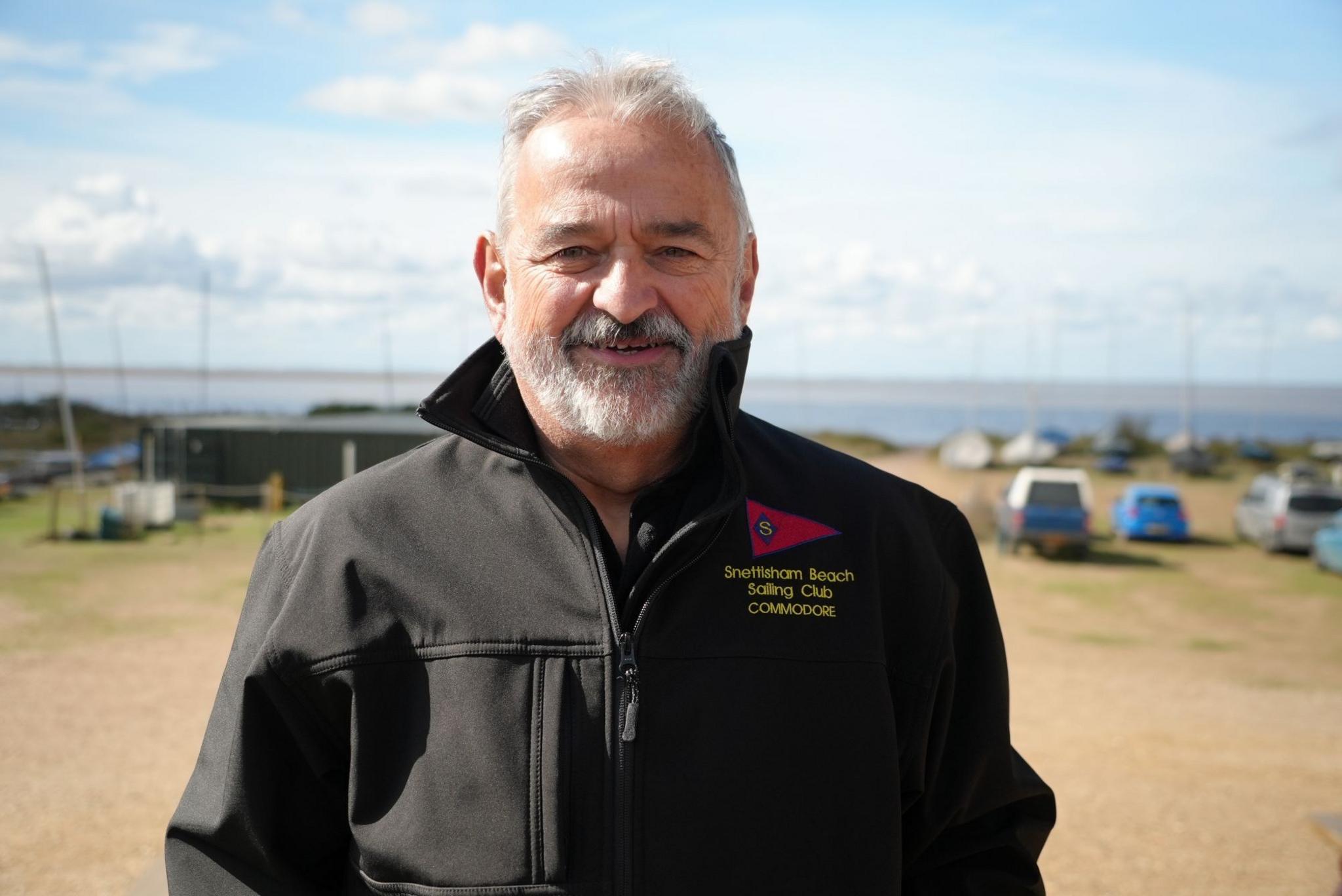 Man with beard in a black jacket. He is smiling and there are boats, cars and the sea in the distance behind him. 