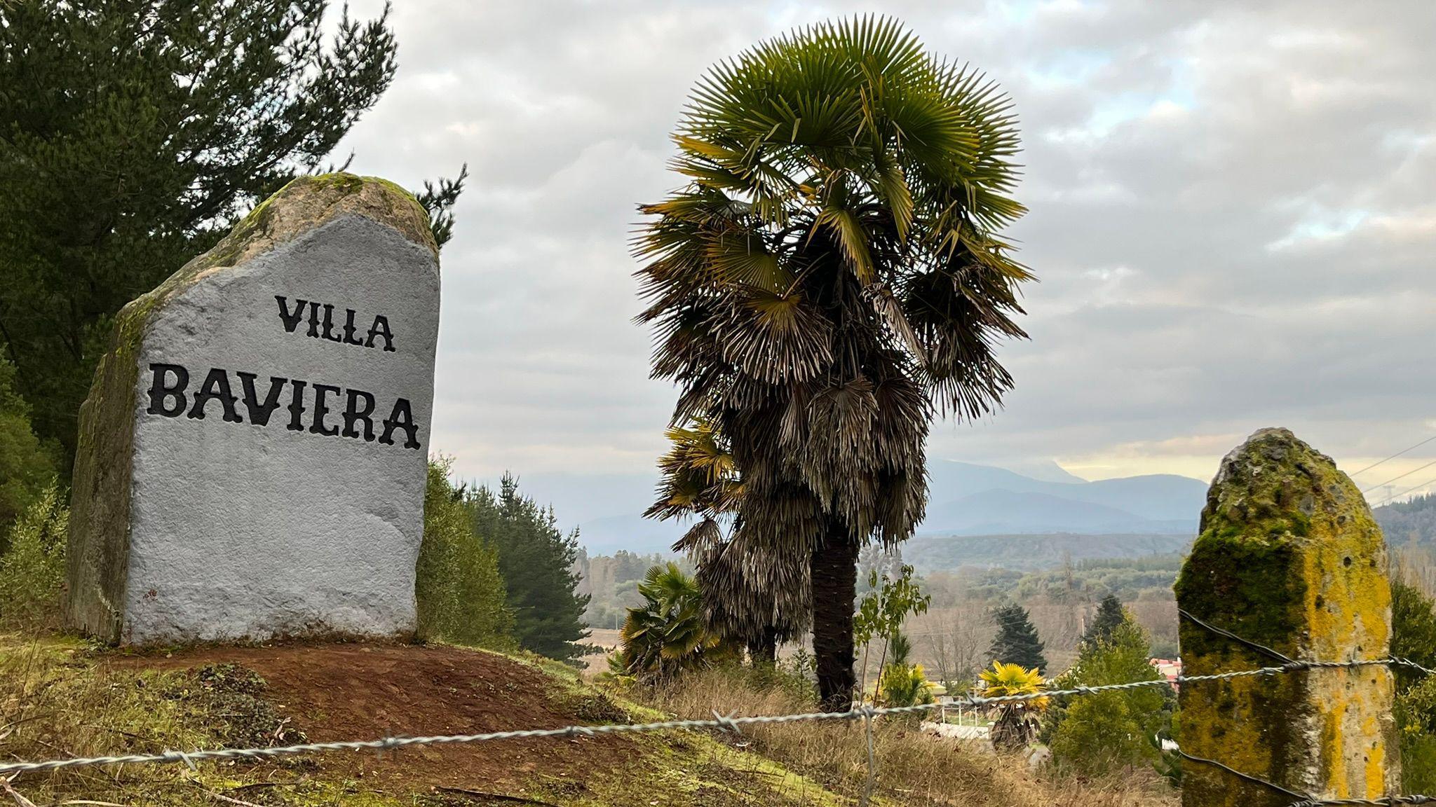 A stone painted white and with the words "Villa Baviera" marks the way to the settlement. A barbed wire fence is seen in the foreground and a palm tree and mountains are in the background