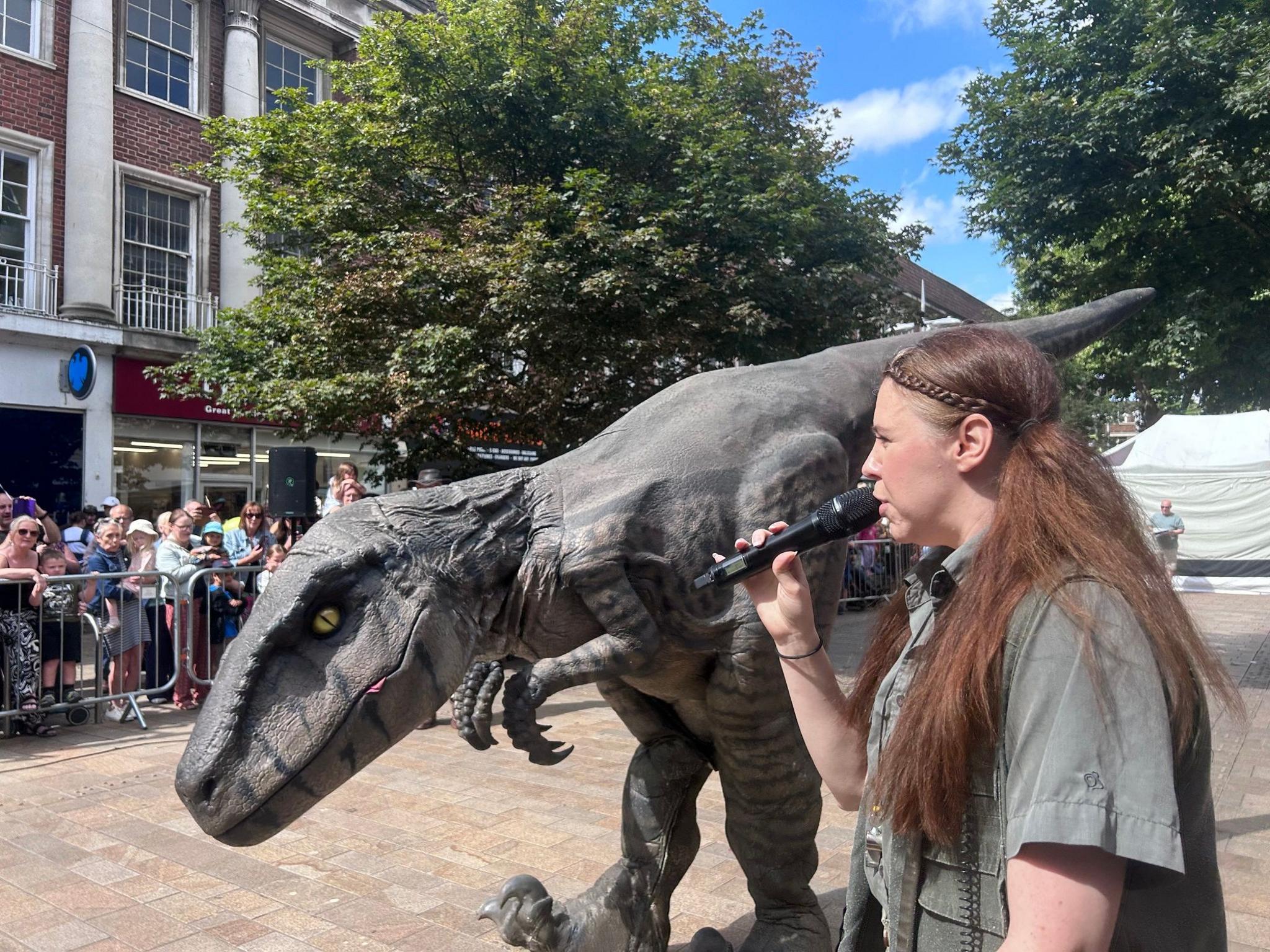A woman with long brown hair wears a grey-green safari-style shirt as she speaks into a microphone while standing close to a large grey dinosaur puppet. They are standing in front of crash barriers and a crowd of spectators, including young children in a shopping street.