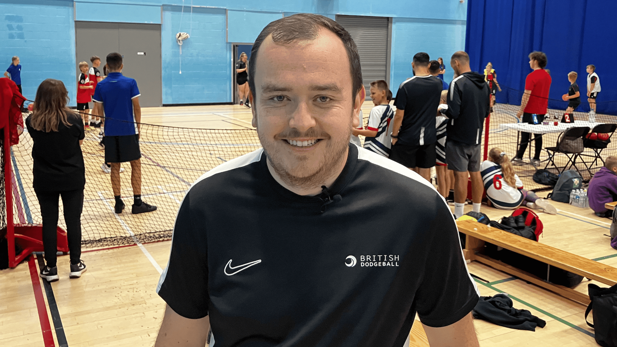 James Robinson from British Dodgeball wearing a black T-shirt. He is standing on a dodgeball court and is smiling into the camera.