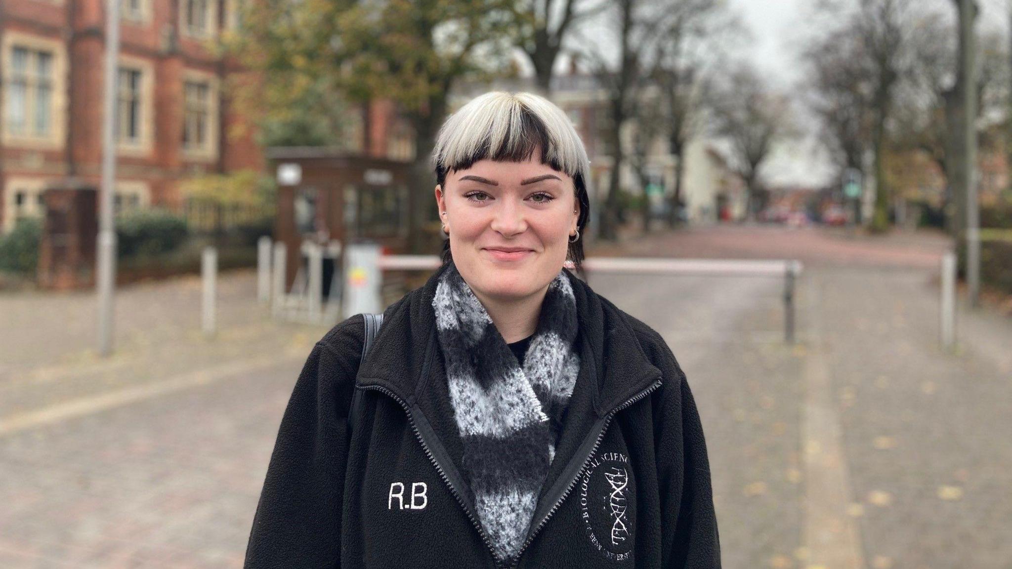 A woman with black and white hair looks at the camera. She is wearing a black jacket and a black and white scarf. In the background there are large red-brick buildings and trees. 