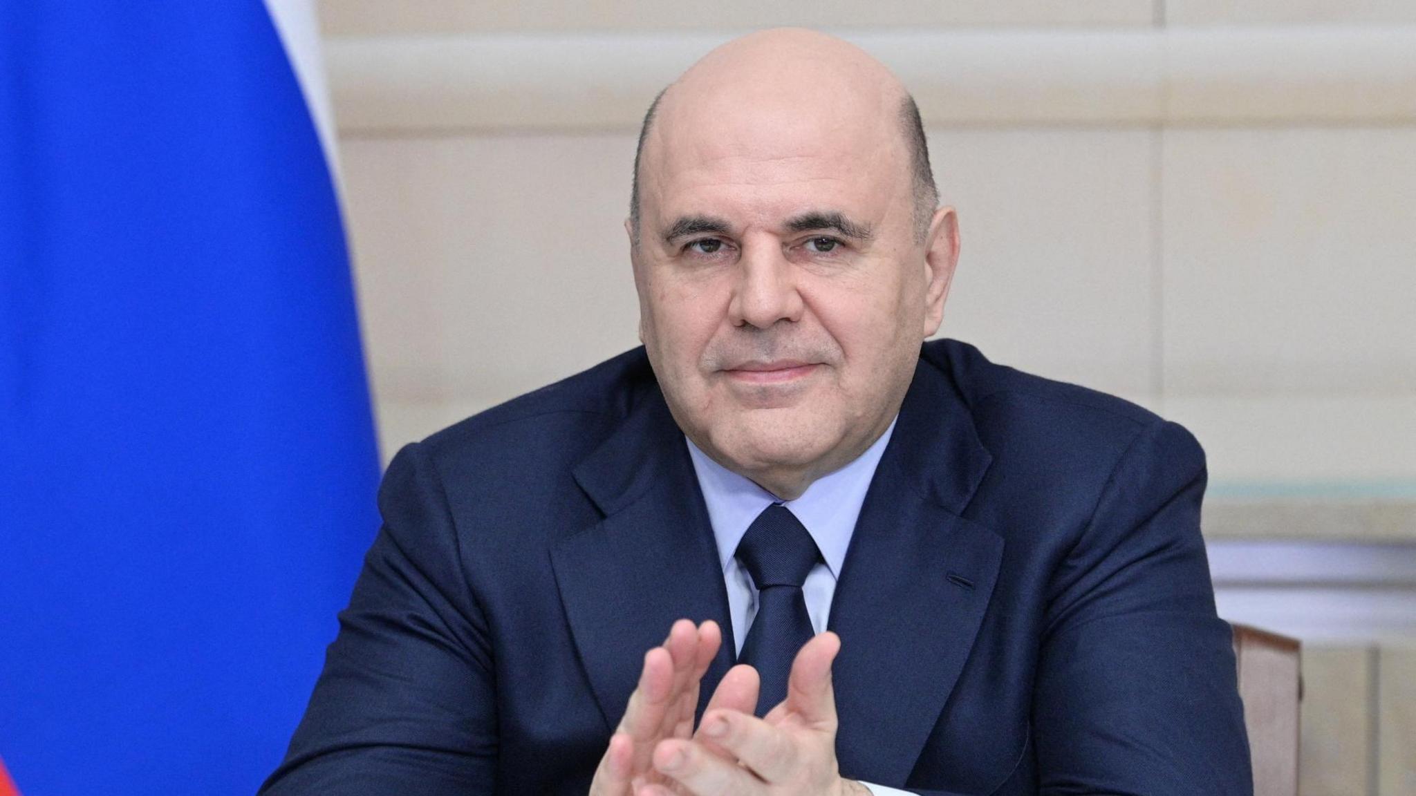 Russian Prime Minister Mikhail Mishustin claps his hands at a ceremony. The blue of a Russian flag falls behind his left shoulder.