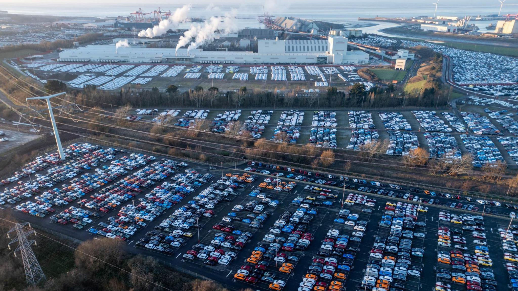 Hundreds of new cars are seen parked up in regimented lines at Portbury Dock in Bristol. The picture is taken from the air on a clear day and the Severn Estuary is visible in the background, as are large industrial buildings, some with steam or smoke coming out of them