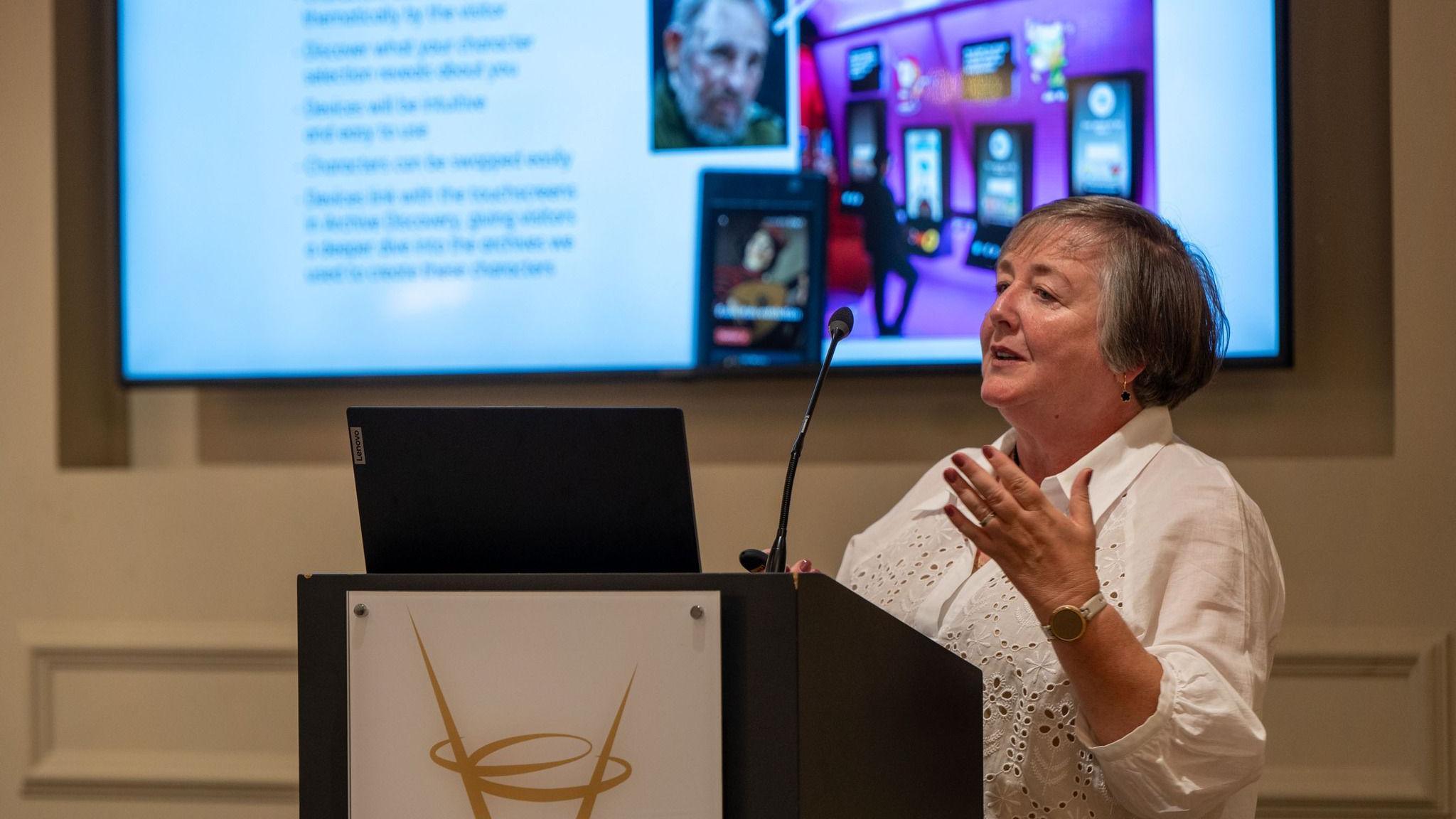 Margaret Edwards, wearing a white blouse and standing behind a lectern, holds one hand in the air. She is speaking into a microphone and standing in front of a large screen