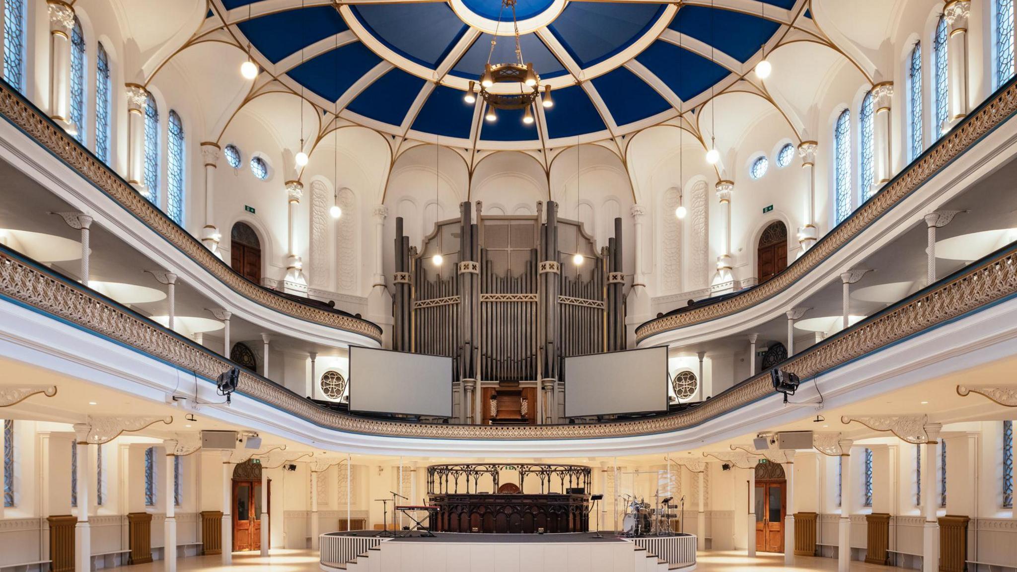 An empty white hall with a large organ in the centre. The ceiling is domed and dark blue. There are two balconies with intricate detailing that run the circumference of the hall. 