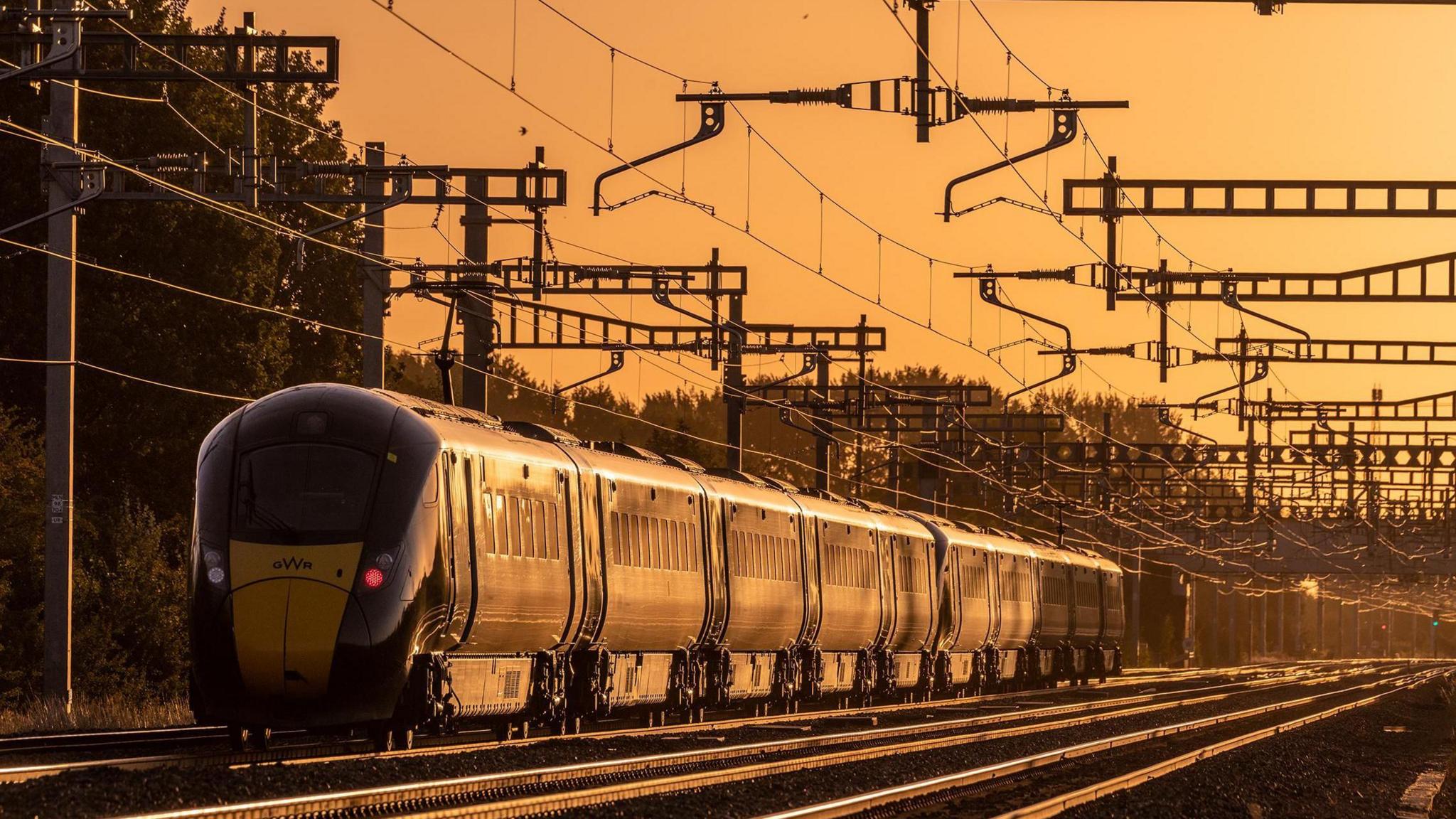 An eight-carriage train in Grove sits in an orange glow created by a low-lying sun and surrounded by overhead power cables