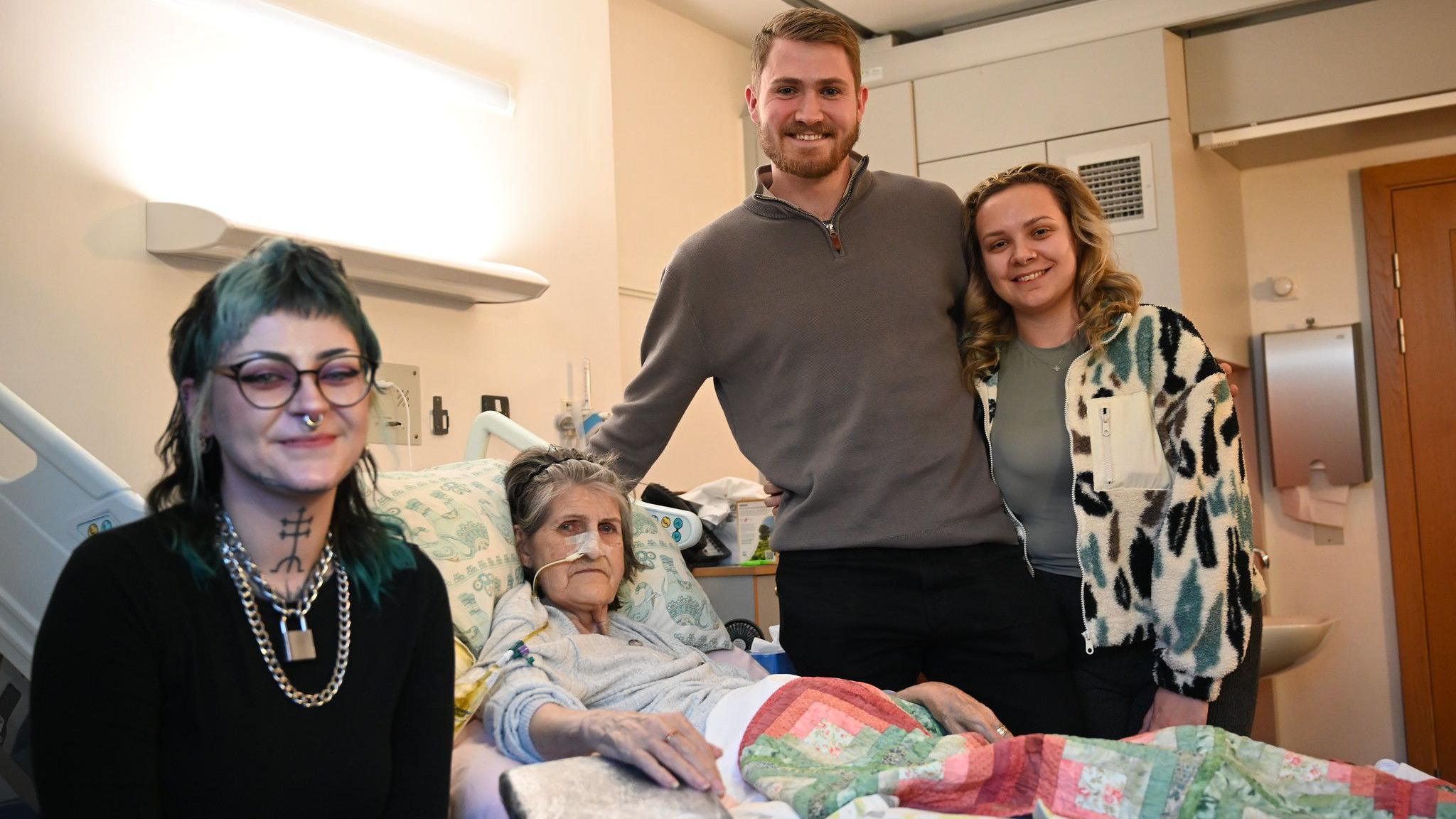 A woman with dyed green and black hair, a neck tattoo, a nose piercing and glasses sits smiling next to an elderly woman in a hospital bed. A young man and a young woman are standing next to the bed, smiling.