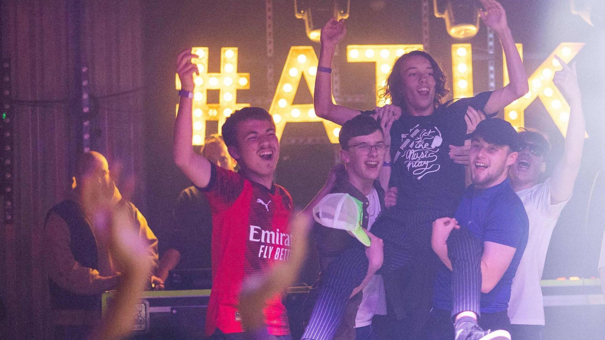 A group of men celebrating and holding the winner in the air holding him by his legs. The men have their arms raised in the air and are smiling and the winner is also raising his arms and smiling at the camera. 