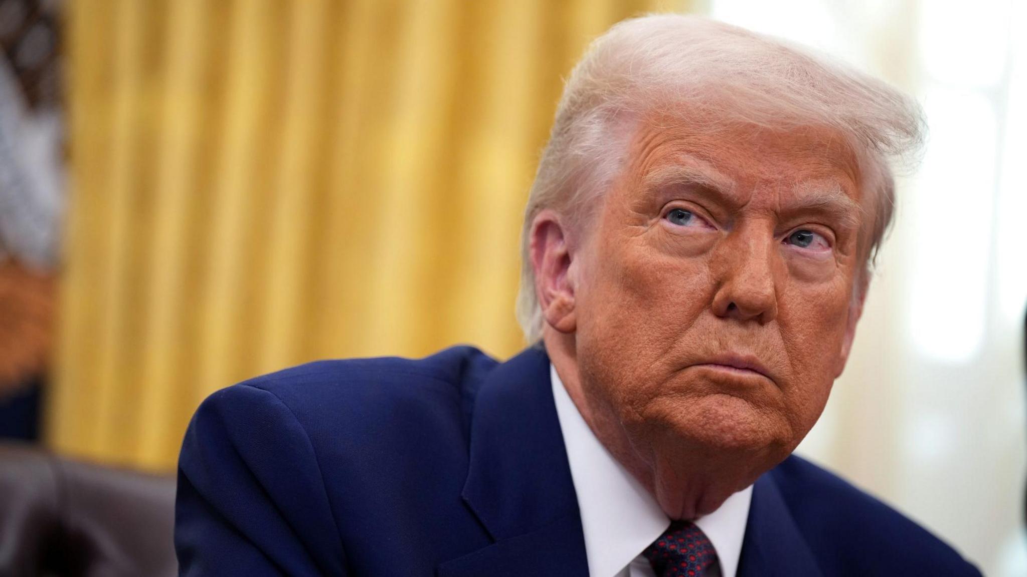 Donald Trump sits at his desk in the Oval Office. He is wearing a navy suit jacket and a white collared shirt and tie.