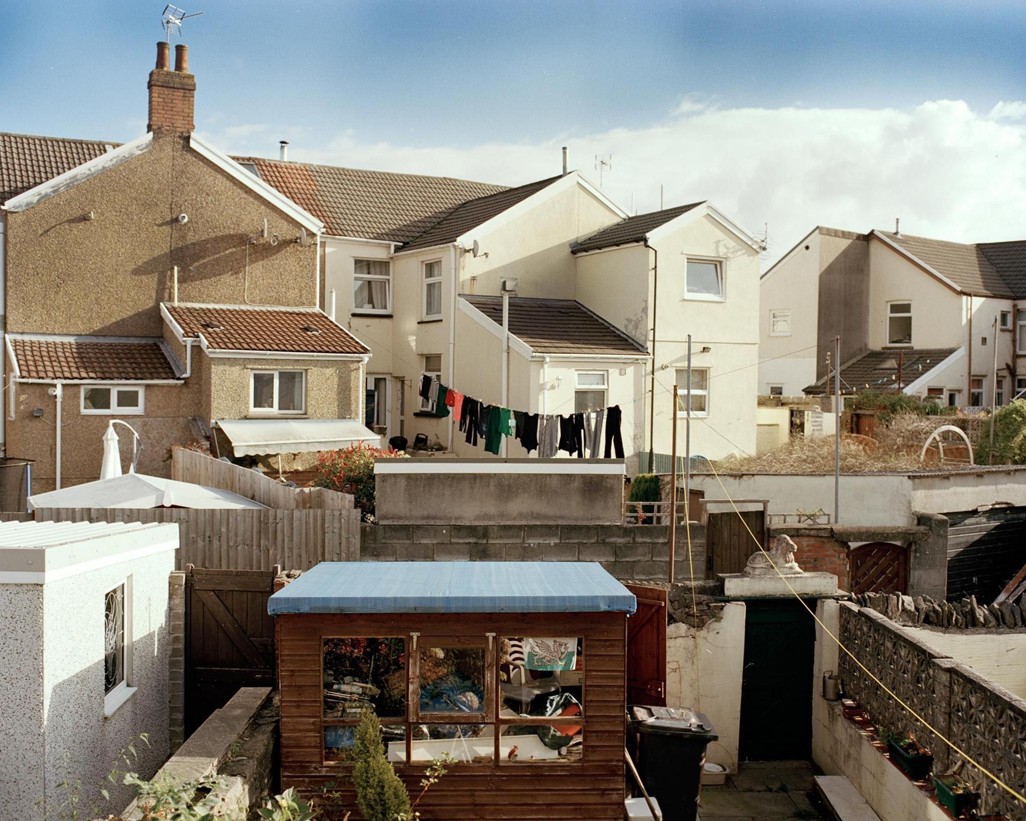 A row of terraced houses photographed from behind with glimpses onto gardens, a shed and a washing line full of clothing 