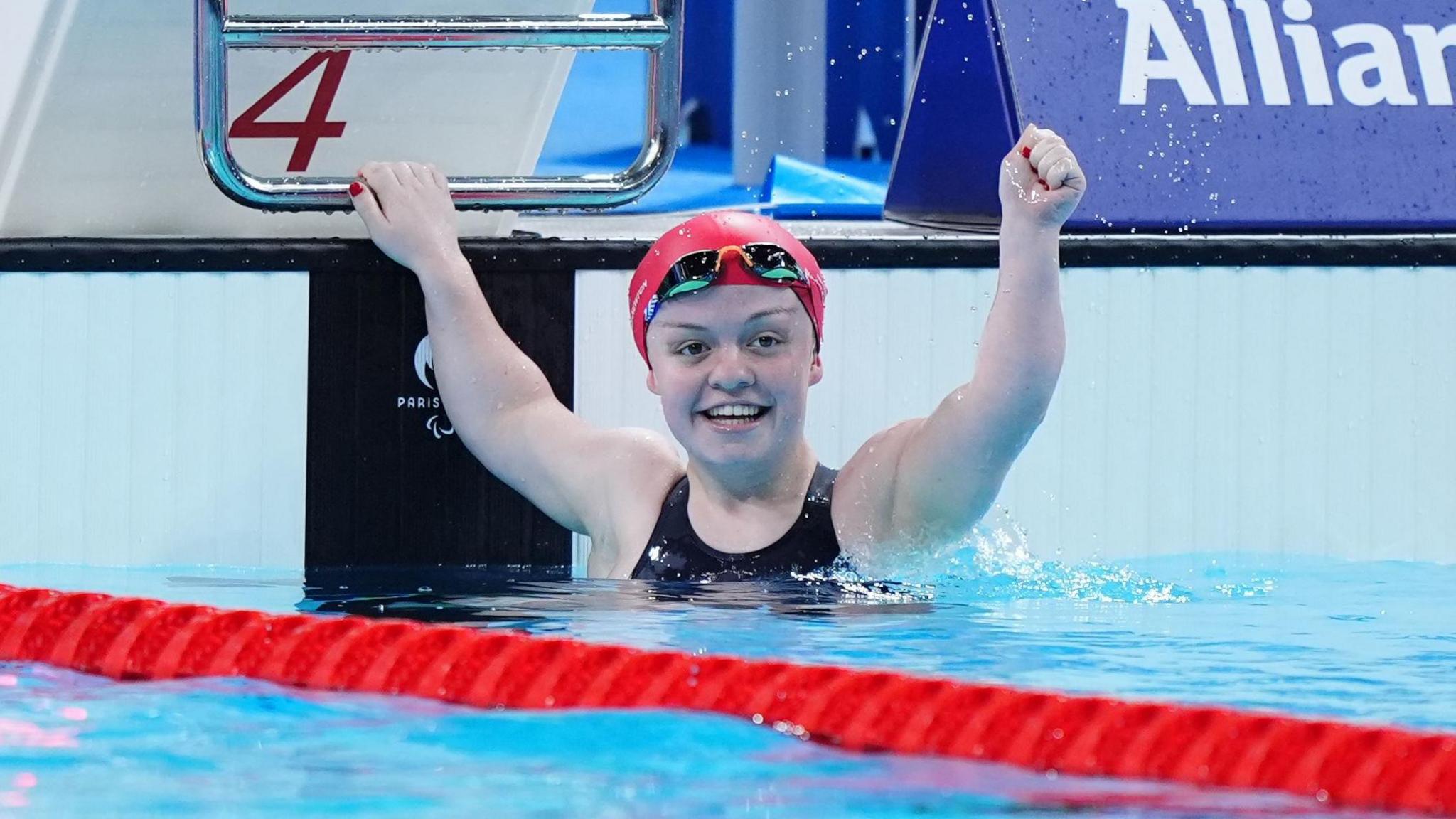 Maisie Summers-Newton, wearing a red cap, black goggles and a black swim suit, lifts her arm in triumph in the pool after winning the Women's 100m Breaststroke - SB6 Final at the Paris La Defense Arena on day four of the Paris 2024 Summer Paralympic Games. 