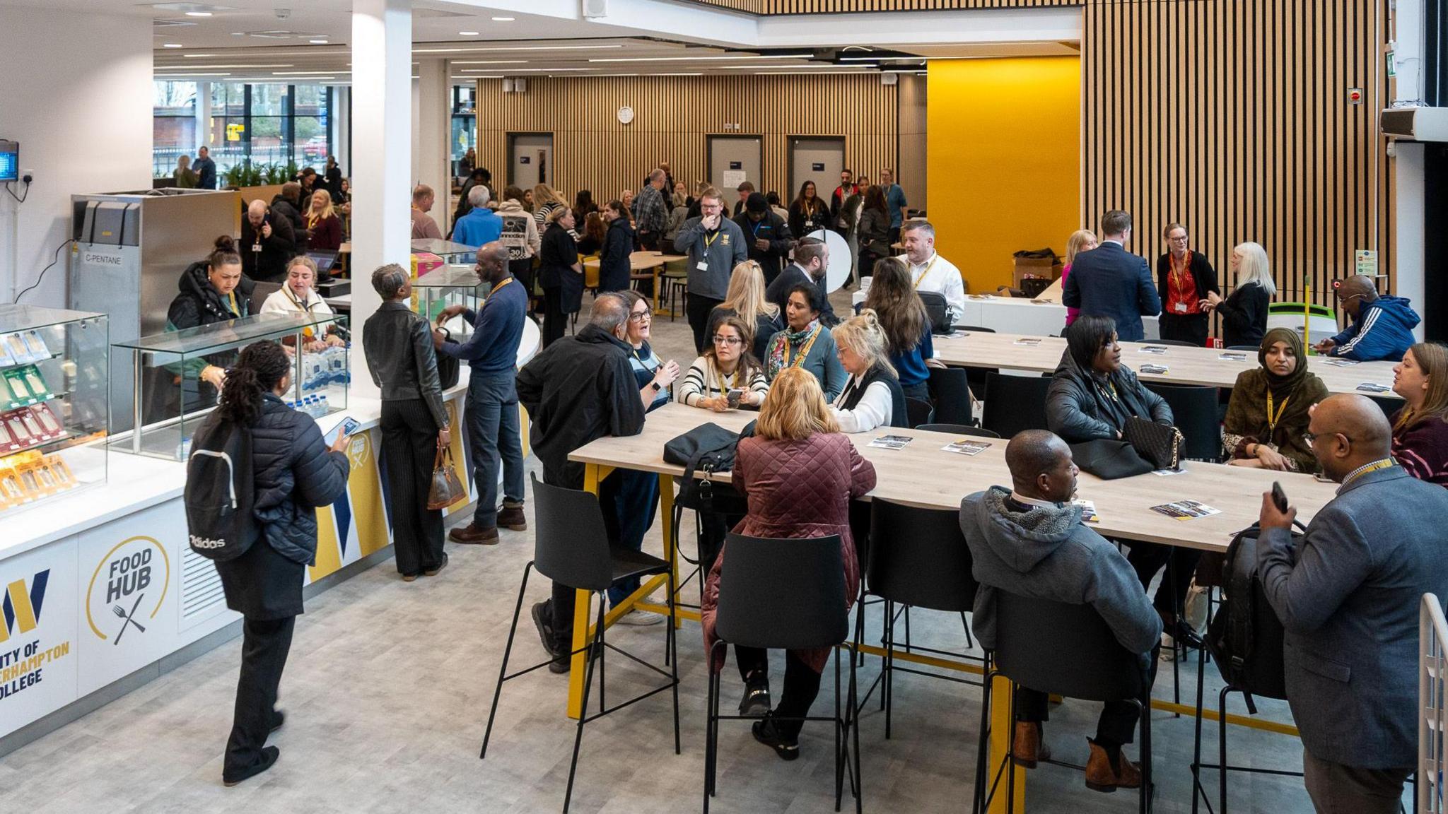A school canteen with a food counter and lots of tables and chairs. The canteen is full with many of the seats taken.