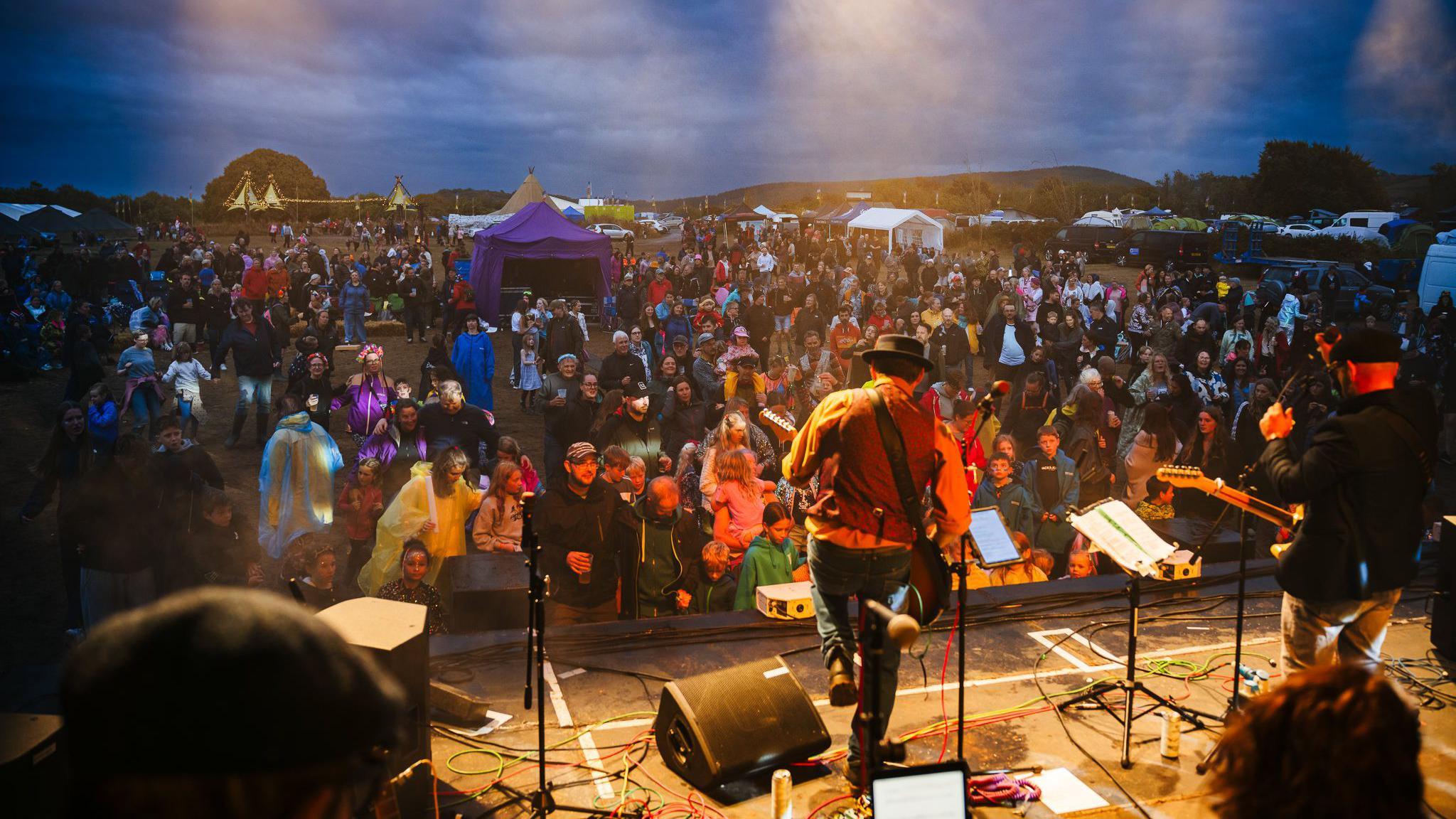 A band called the Jamestown Brothers perform on stage at the Mendip Family Festival. The picture is taken from the back of the stage with an audience of hundreds of people visible. Small tents and food stalls are visible around the edge of the auditorium