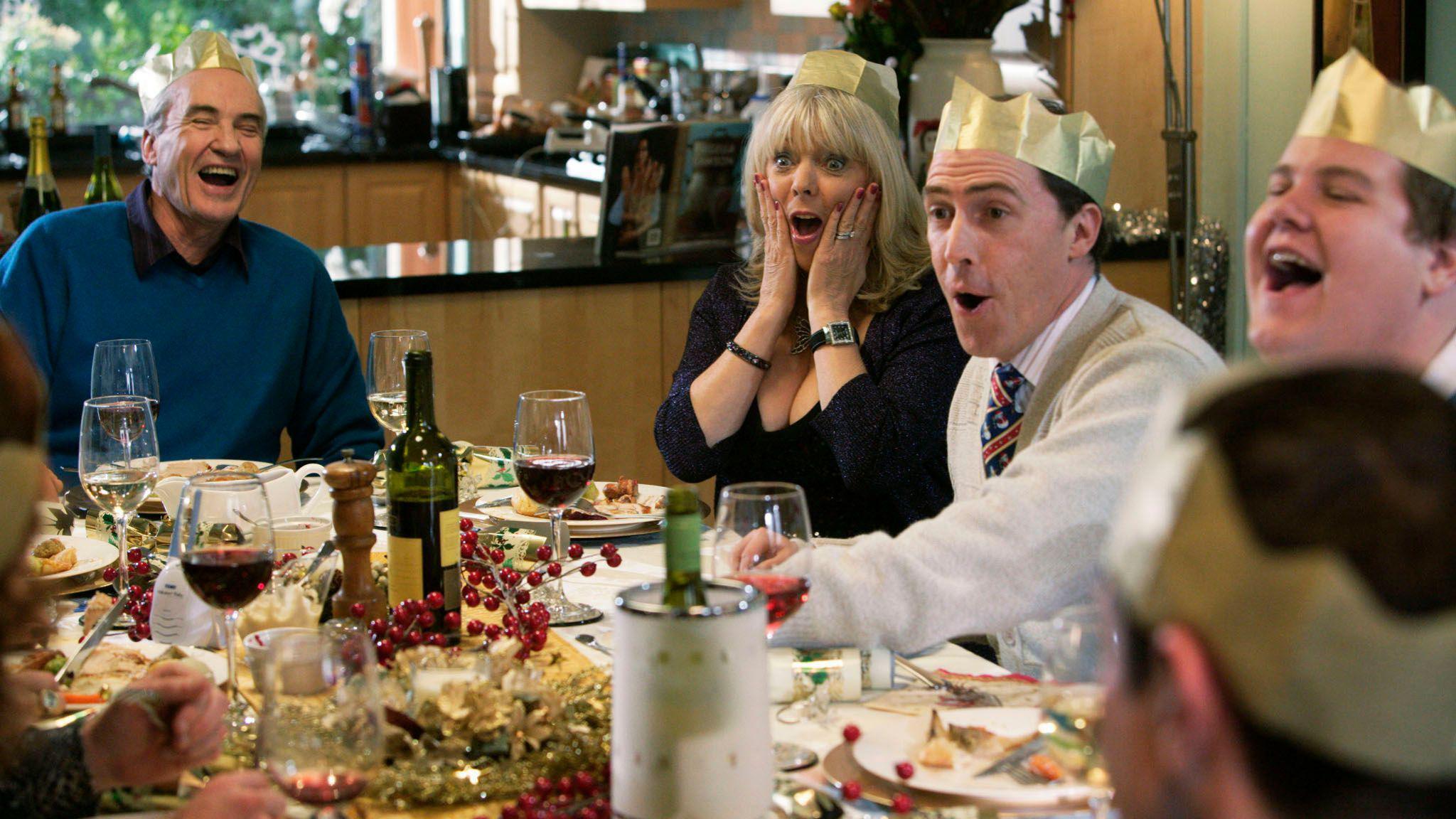 The cast of Gavin and Stacey sitting around a dinner table at Christmas. Everyone is wearing gold paper crowns and laughing. On the table there are plates full of roast dinner, Christmas decorations and glasses of wine. 