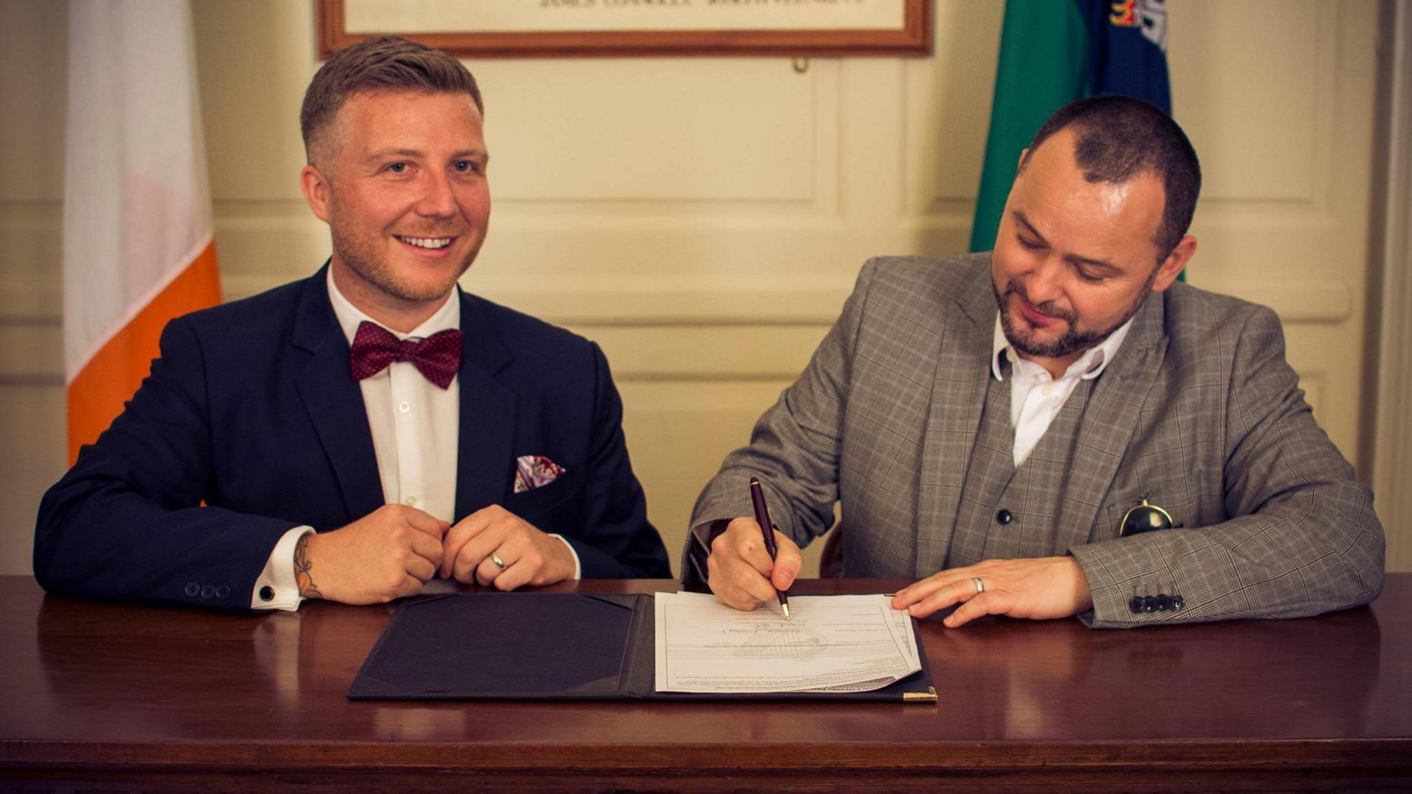 Two men signing marriage documents, one is wearing a navy suit. The other is wearing a check grey suit.