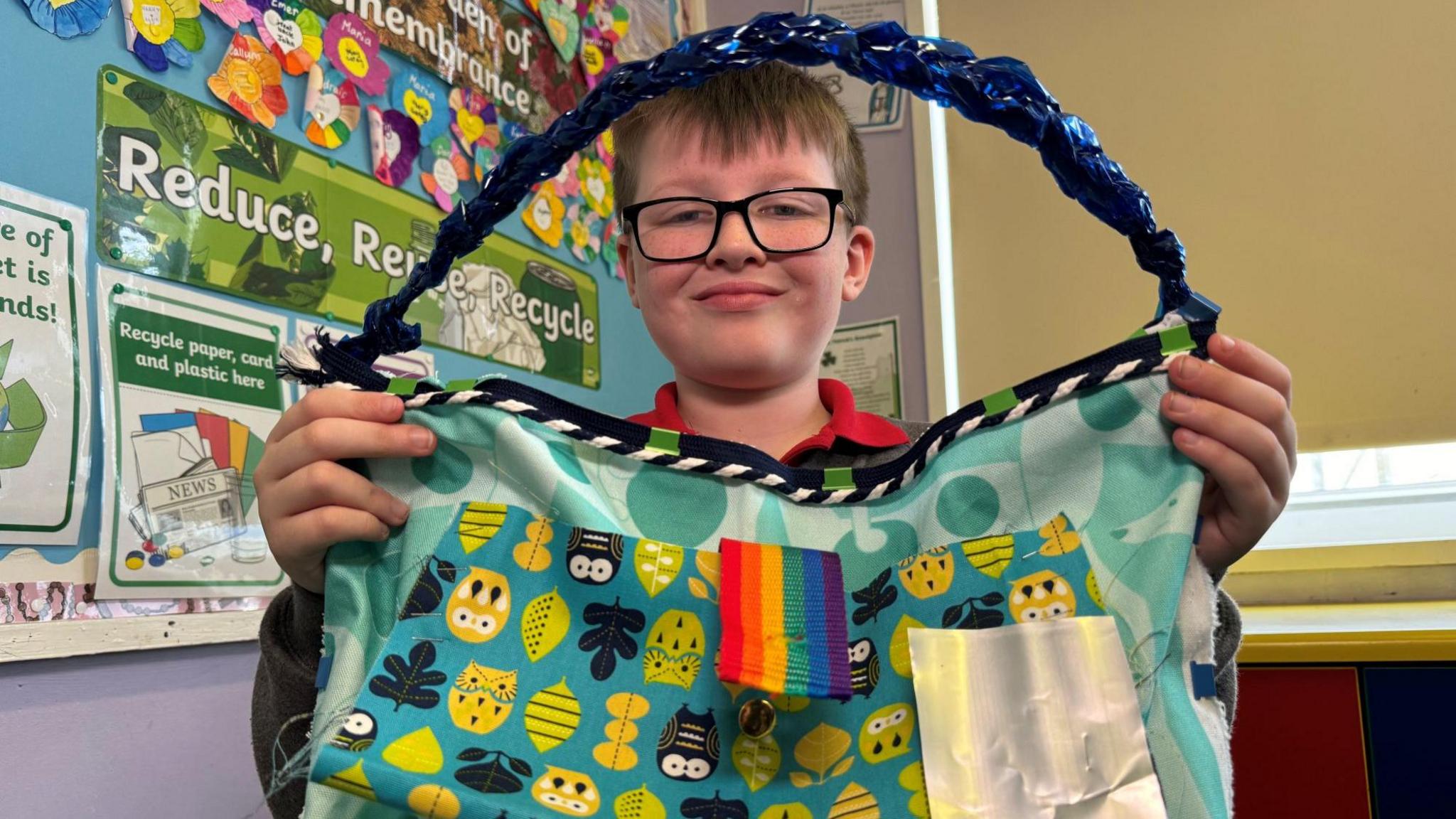 Cillian - a young child wearing black, square framed glasses holds up a large blue, leaf patterned bag with a blue, plaited handle. 