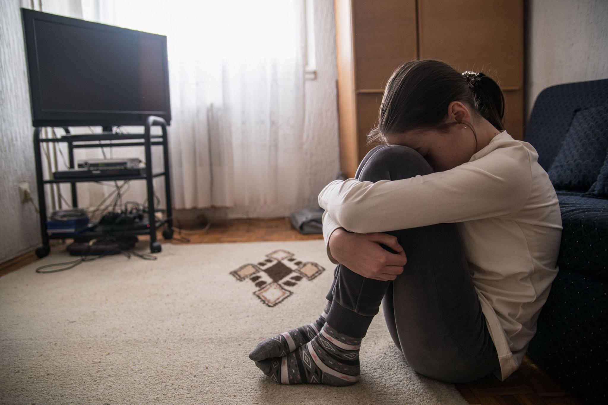 A young girl appears sad as she puts her head on her knees in a small flat. 