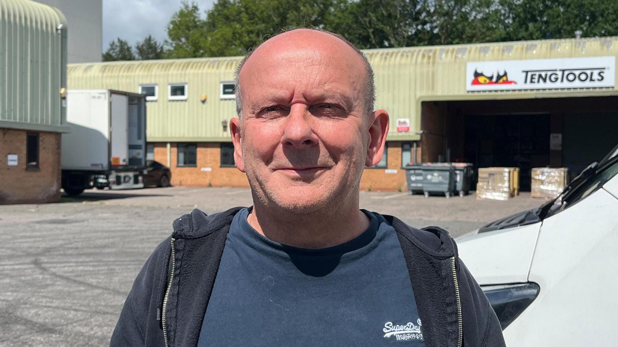 A bald headed man wearing a blue T-shirt and zip up top smiling into the camera. He is standing in an industrial estate with a van and tool company in the background.