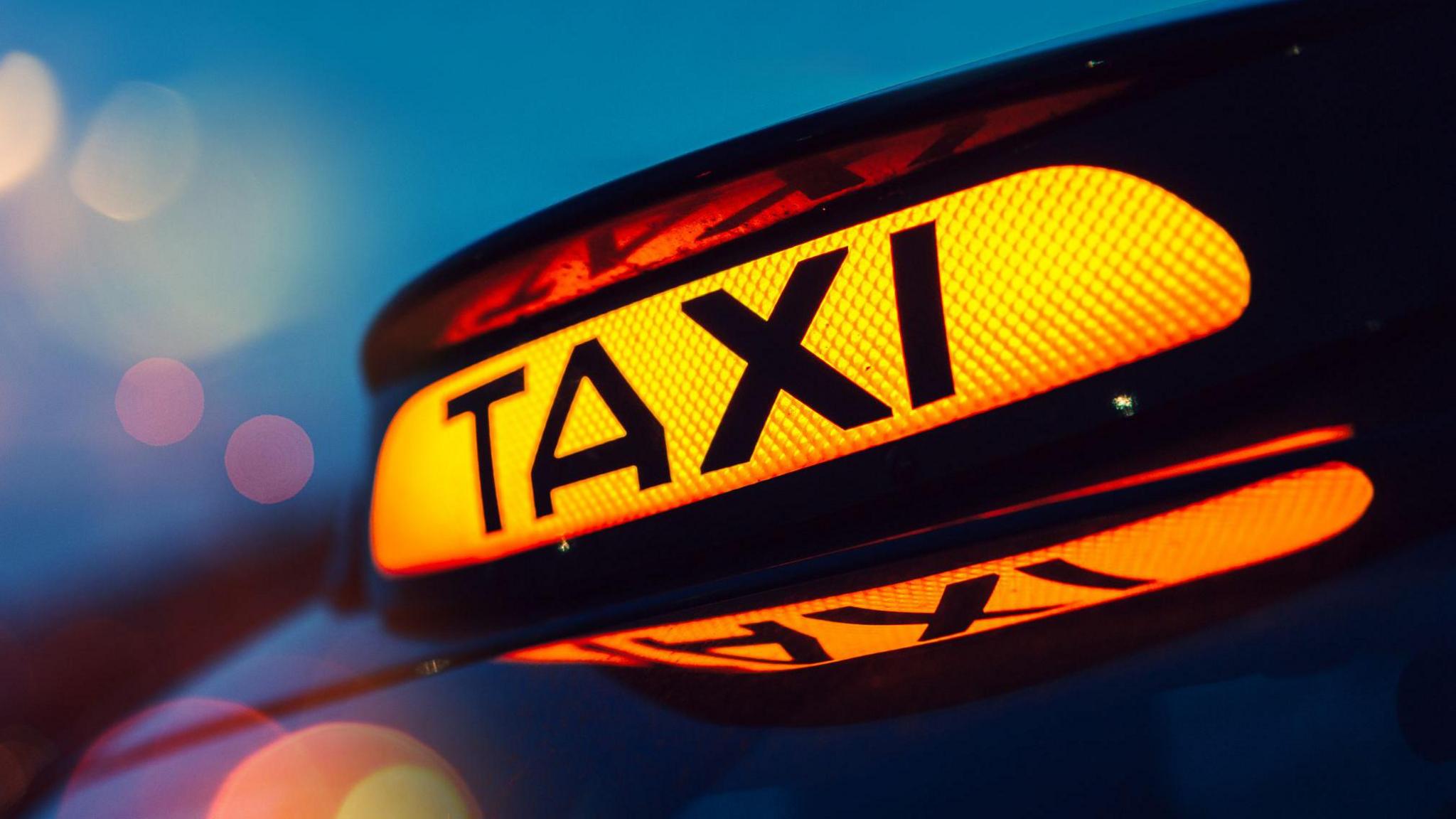 Close up of an illuminated taxi sign. It is a yellow light with the word taxi written across it in black letters