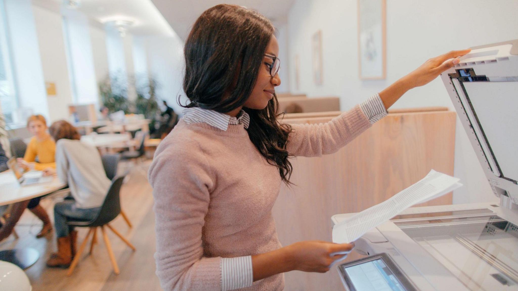 Photo of a young woman in an office using a photocopier while her co-workers are having a meeting behind.