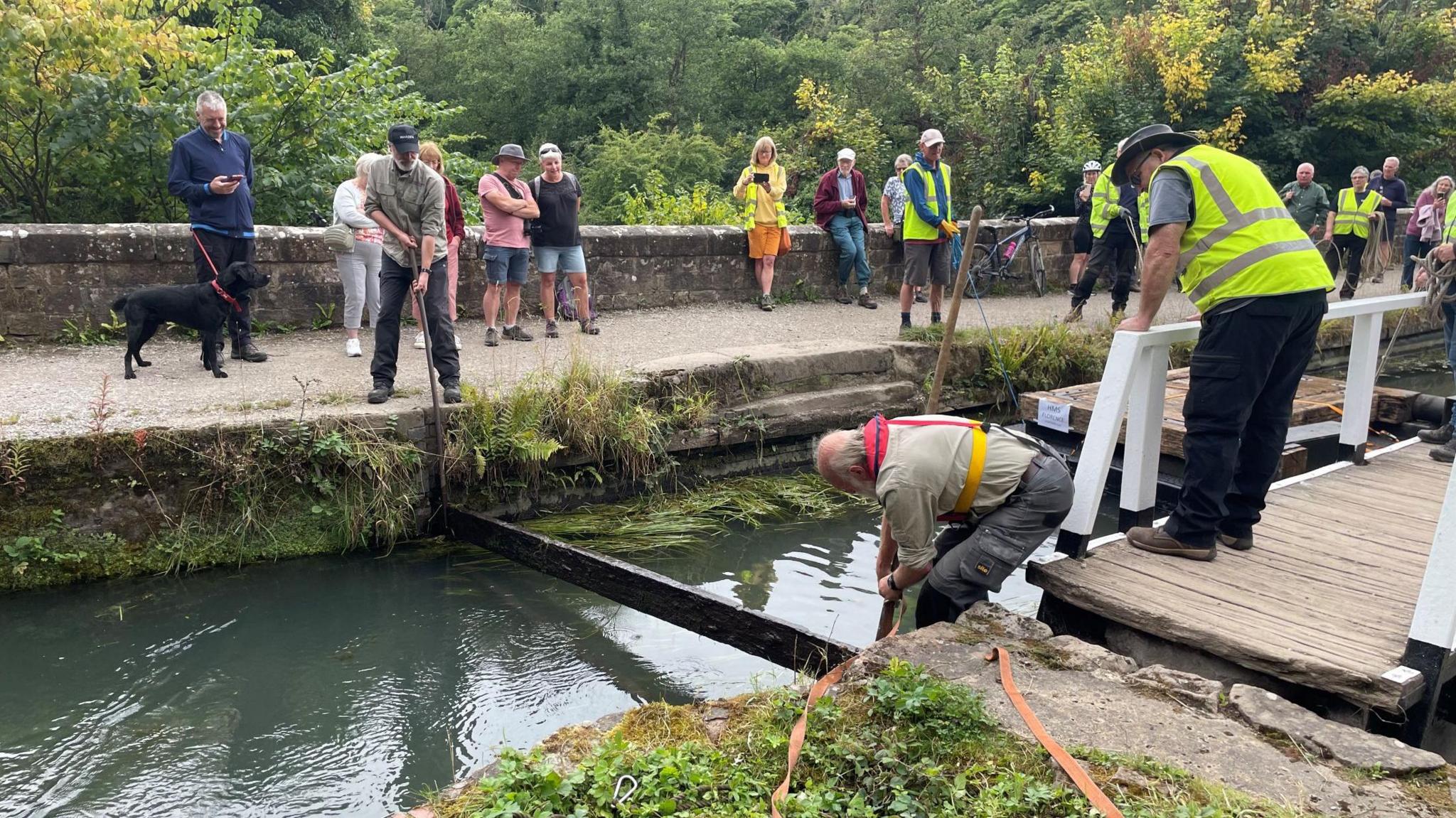 People watching the journey of the lock gate on the canal, as a volunteer lifts a stop plank out of place for the pontoon to pass 