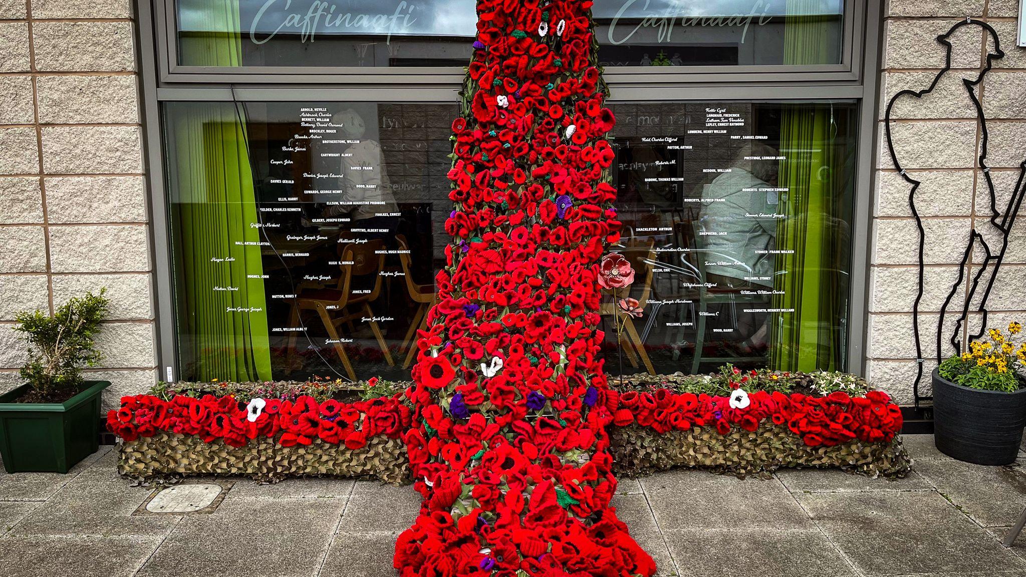 Red poppies from a five metre long cross that stretches up the window of a building. Beside the window, on the grey brick wall is a black wire outline of a soldier.
