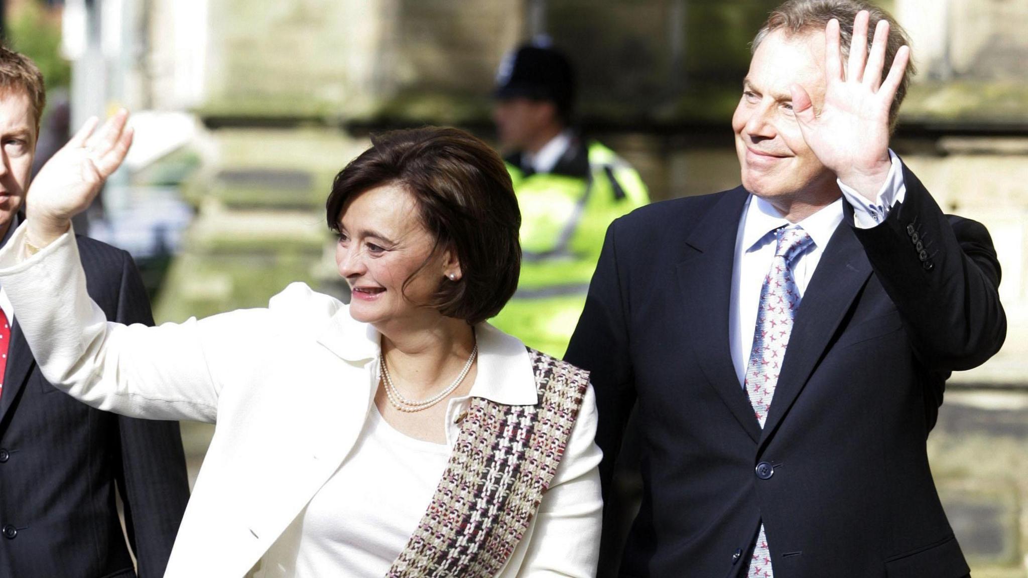 Cherie and Tony Blair wave and smile. She has short dark hair and is wearing a cream suit and top with a Burberry-pattern scarf hung over her left shoulder. He is wearing a dark suit and a silver tie with a red pattern