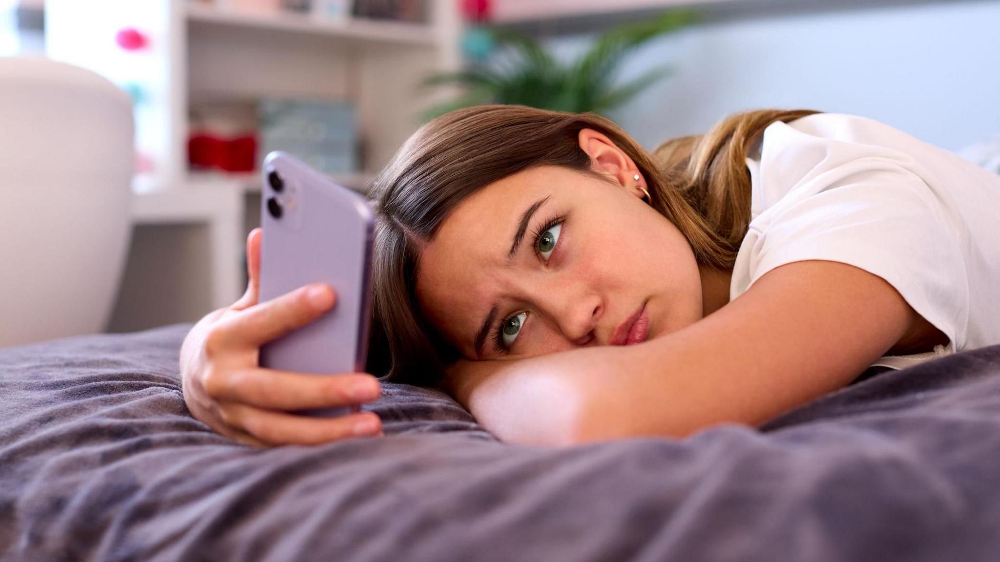 A teenage girl is lying on a bed staring at her phone which is in a purple case. She looks concerned. the bedding is purple.