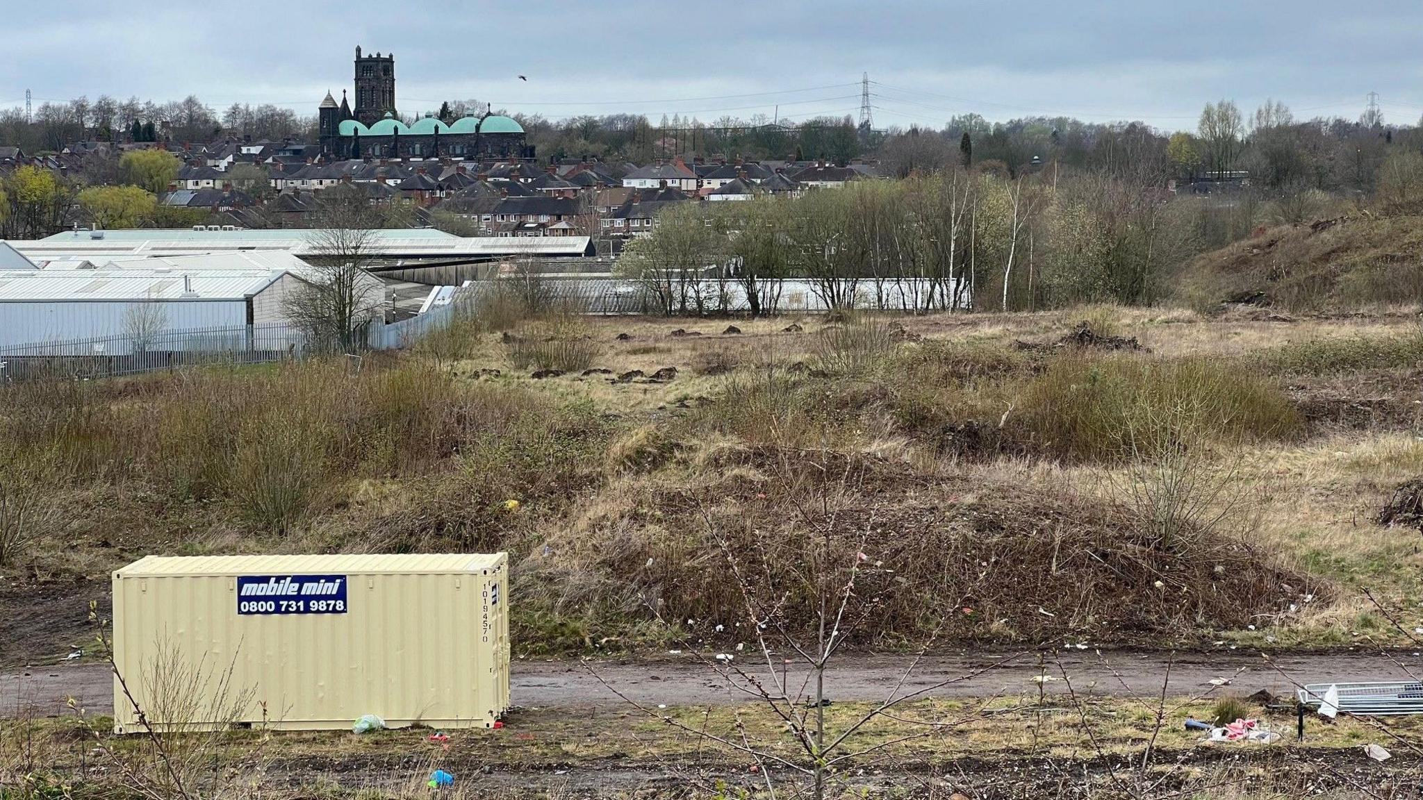 A shipping container in the foreground, sitting on a patch of scrubland, with a industrial units and a town centre in the distance.