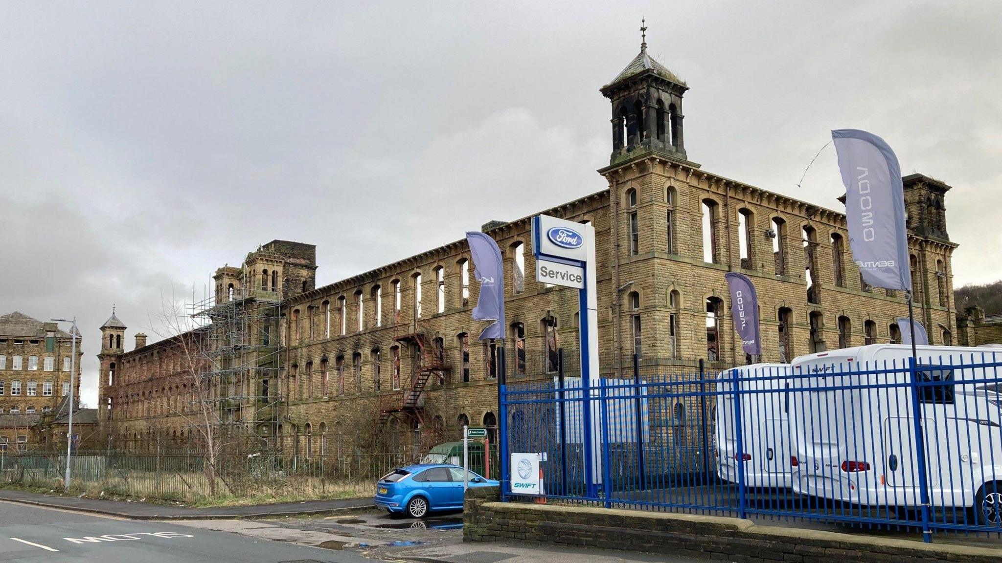 The Yorkshire stone shell of a former four-storey mill with scaffolding running up two towers and daylight clearly visible through its window arches