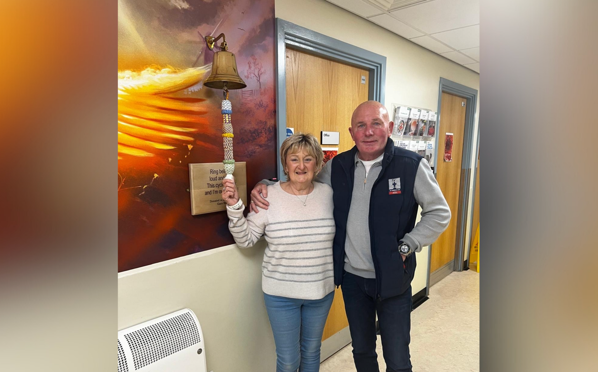 Joan, standing next to her partner Ron, holds on to the bottom of a rope which is attached to a gold coloured bell at Newcastle Freeman Hospital. It signifies the end of her treatment following her breast cancer diagnosis.