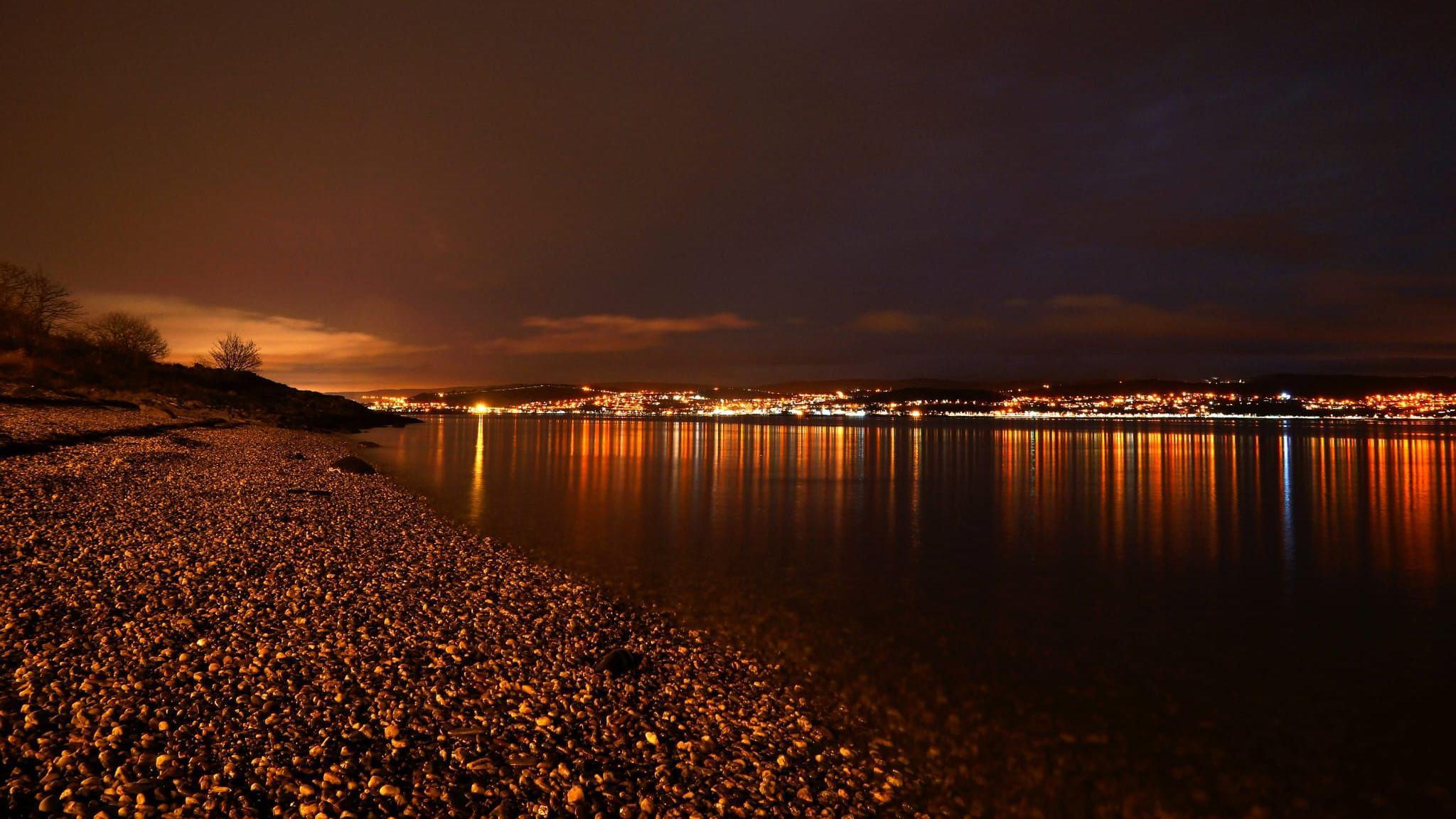 A night scene of the lights of a town glowing orange across a body of water.