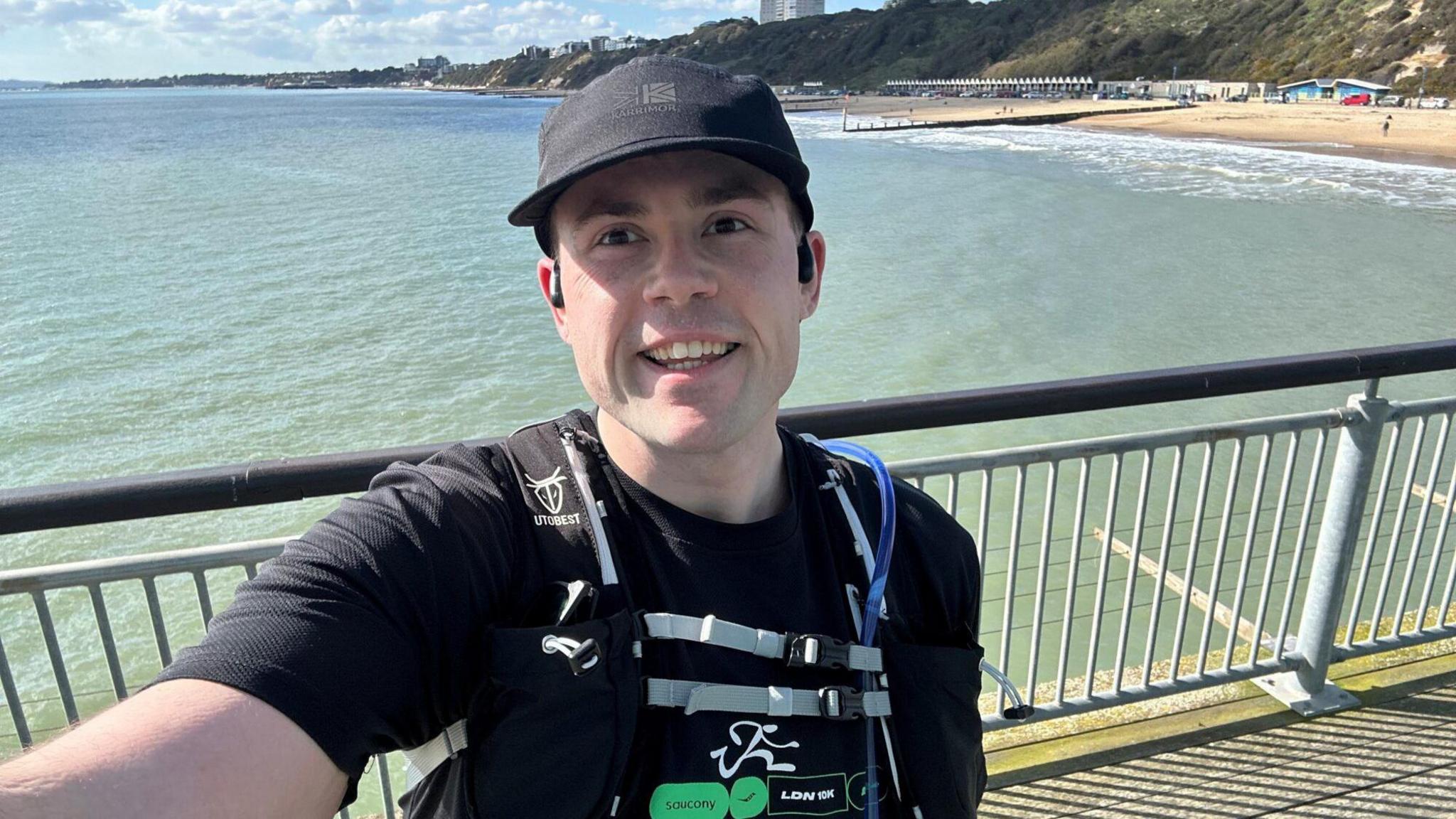 A man in a blue top with a backpack. He is stood in front of railings and smiling in a selfie picture.  There is a beach behind him.