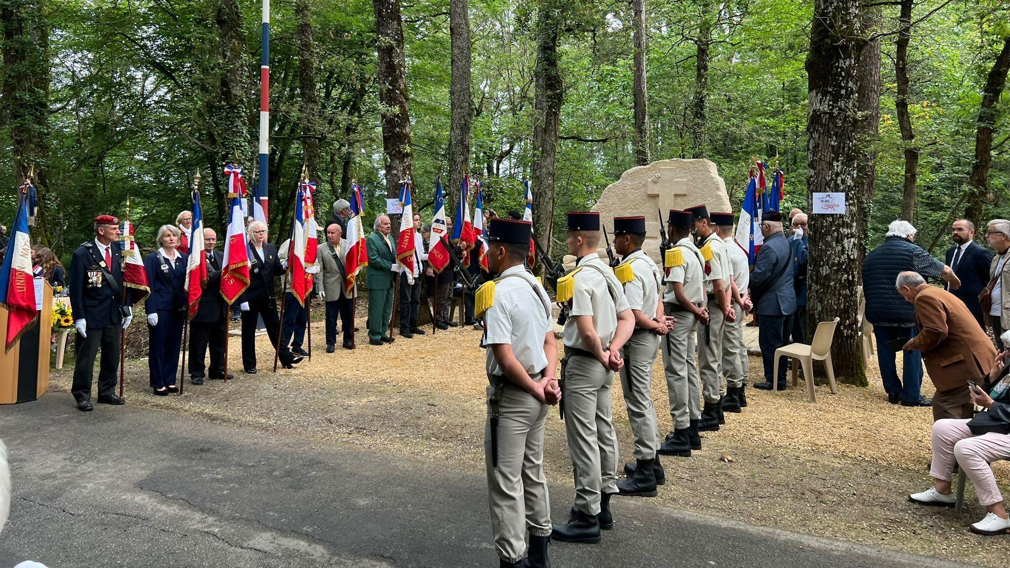 Representatives of the French army and veterans' organisations stand in two rows by a large stone monument in a forest