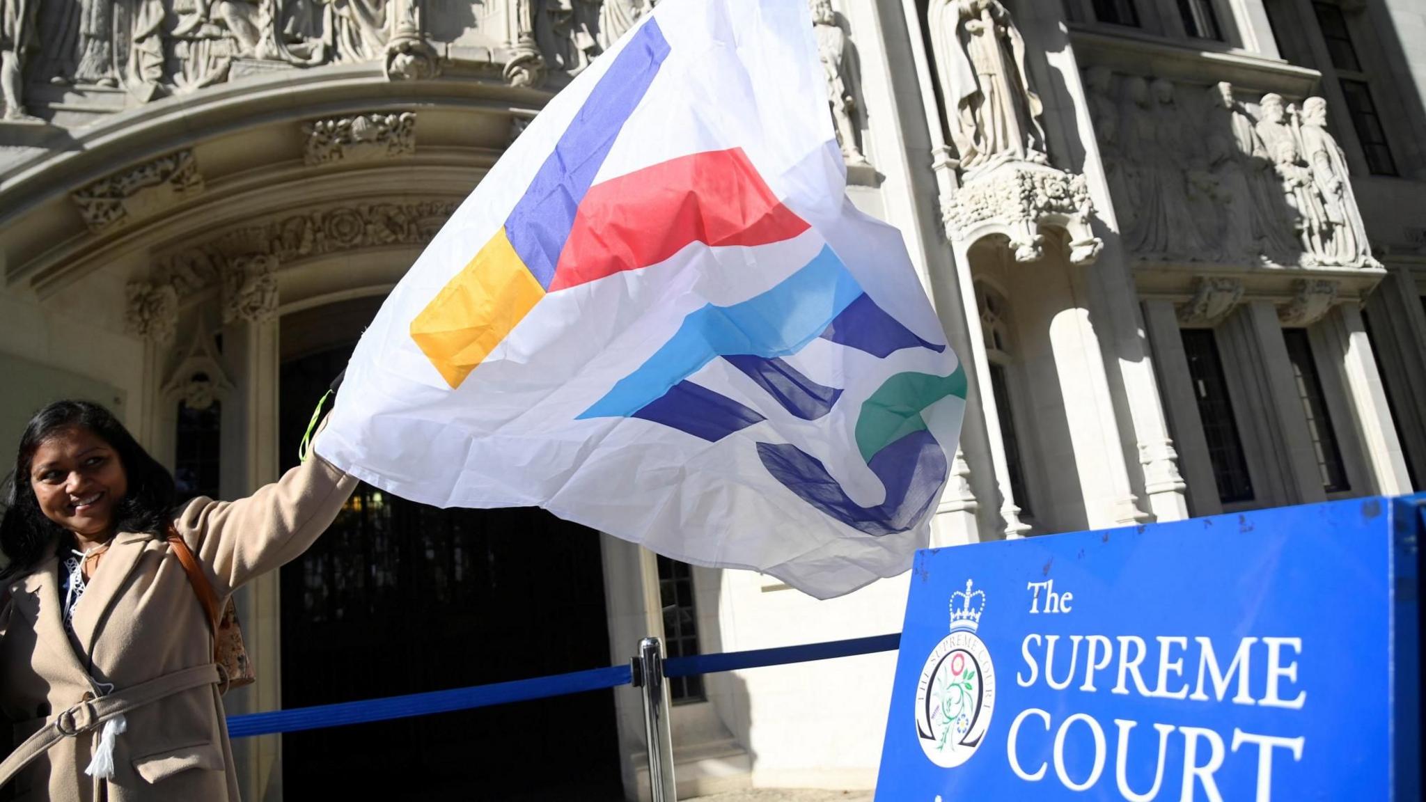 A female pro-Scottish independence campaigner in a beige coat waves a white flag which spells out Yes in multi-coloured letters. She is standing outside the UK Supreme Court building and there is a blue court sign to the right.