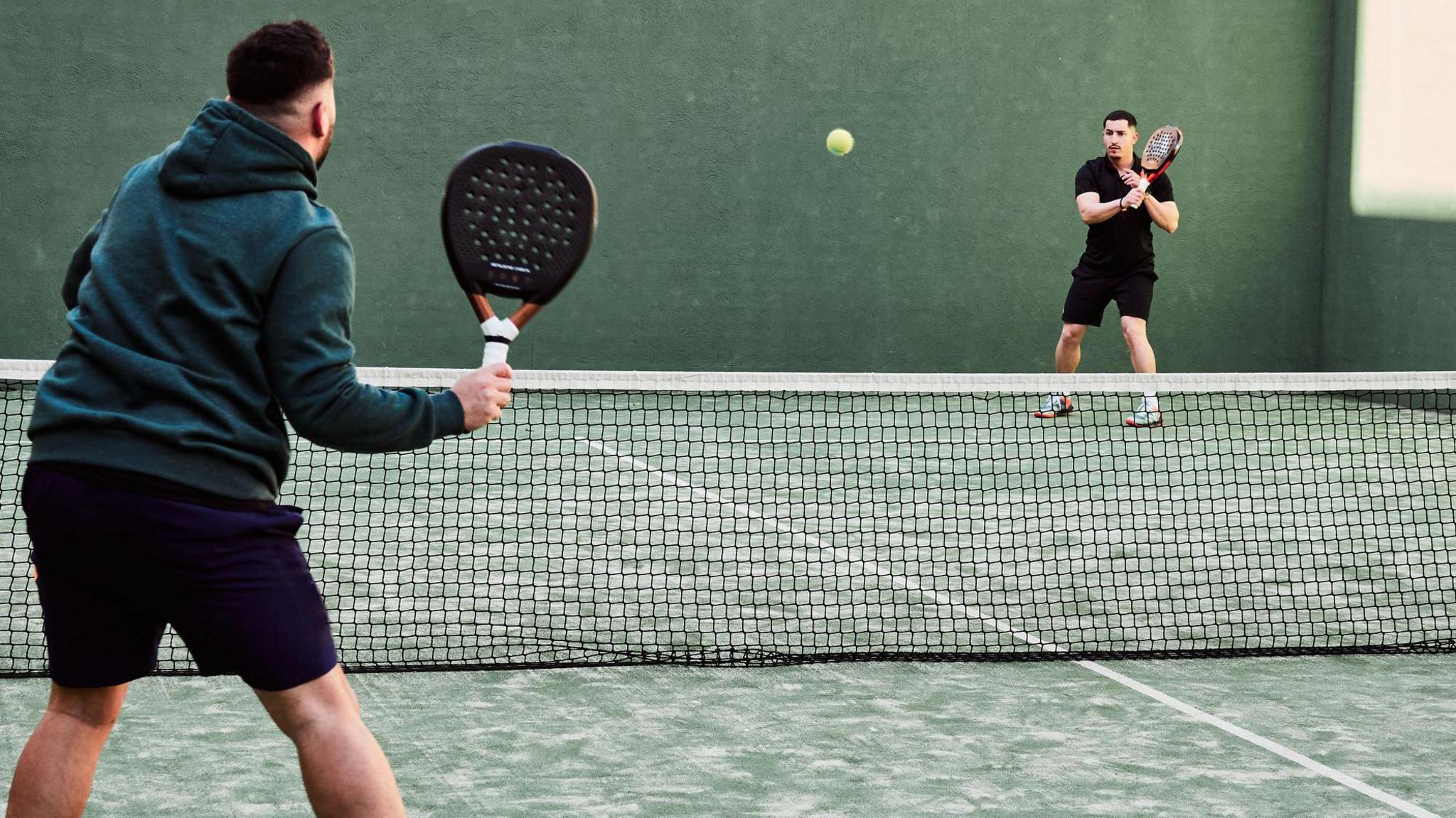 Two men playing padel on an outdoor court, with one preparing to return a shot while the other stands near the net.