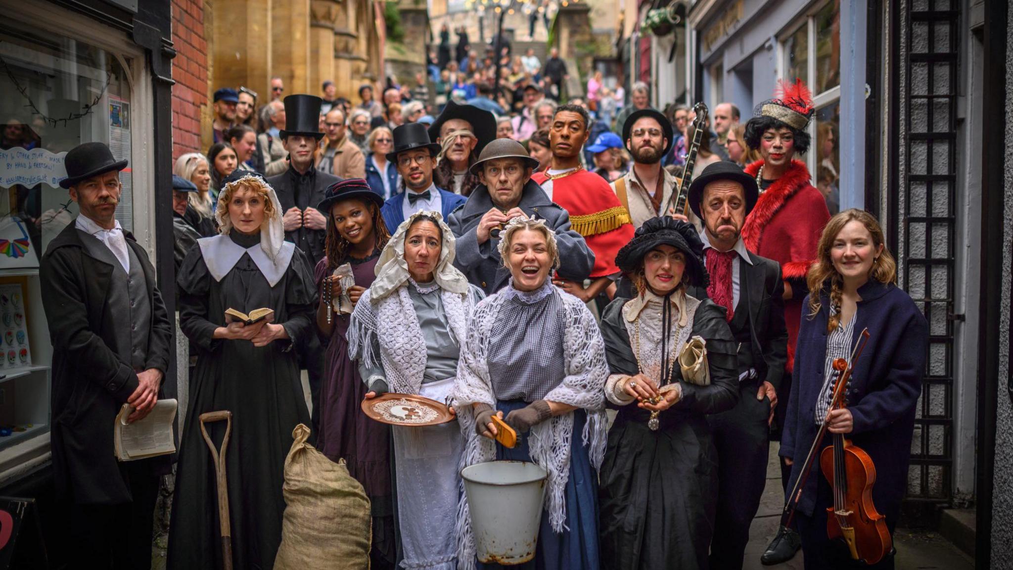 A large group of performers in various old-fashioned outfits smile at the camera as they stand on the Christmas Steps in Bristol