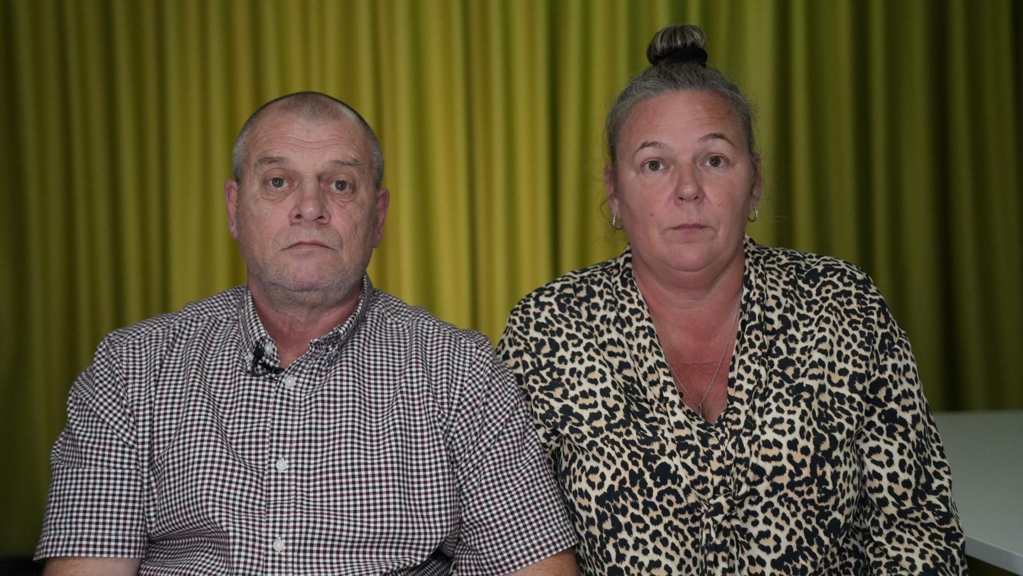 Ray and Collette Kerry sat next to each other. Ray has a shaved head and is wearing a black and white chequered shirt. Collette is wearing a leopard print top and has her grey hair tied in a bun. Both are serious in their expression and sat in front of yellow curtains.