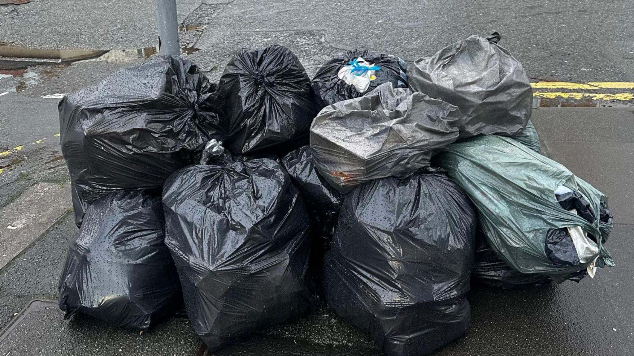 Ten full black and grey bin bags lie in a pile on a street corner next to a lamppost