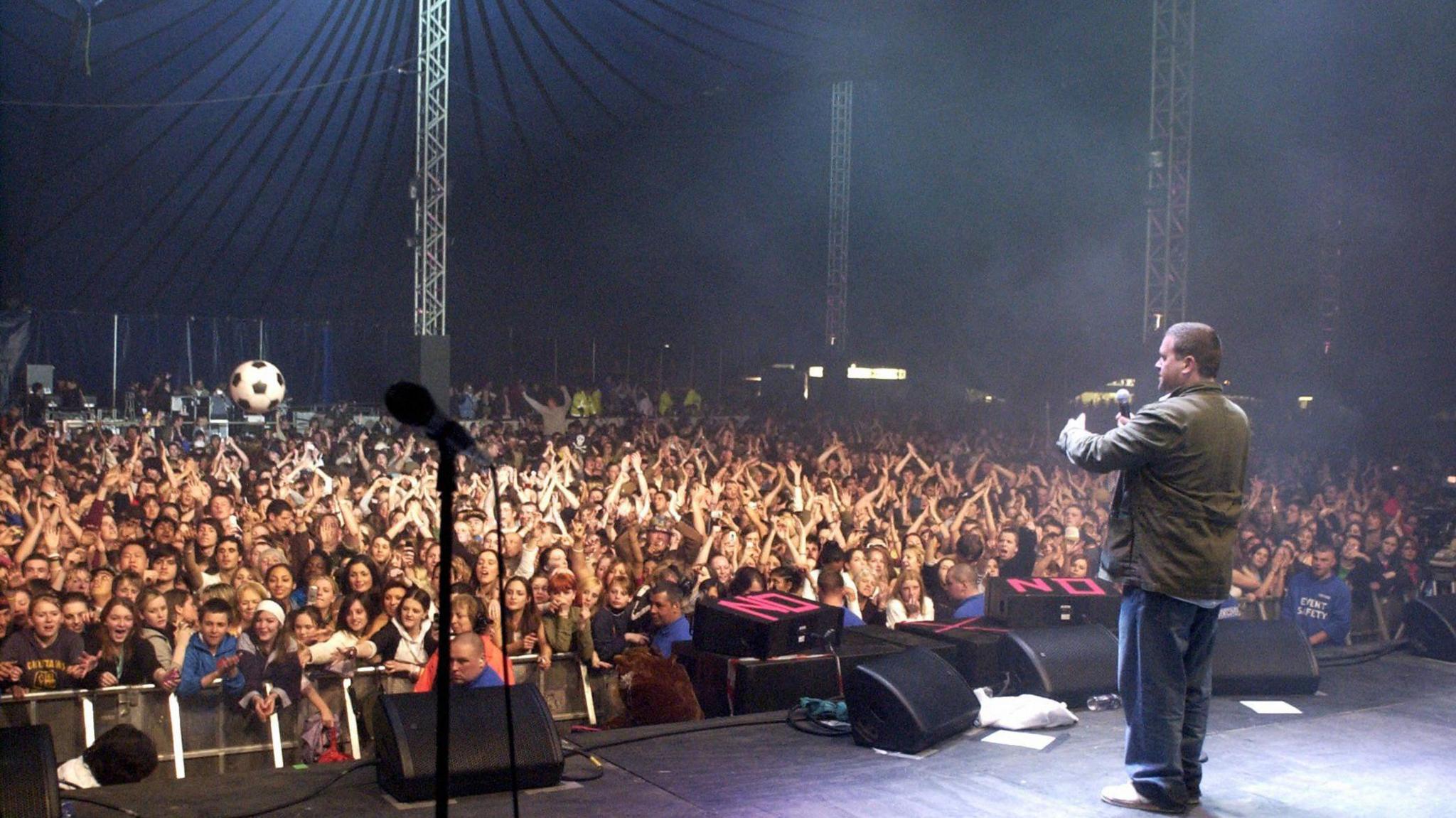 A large crowd of people inside a giant blue marquee watches and applauds as DJ Chris Moyles, who had short dark hair and is wearing blue jeans and a green jacket, addresses them.